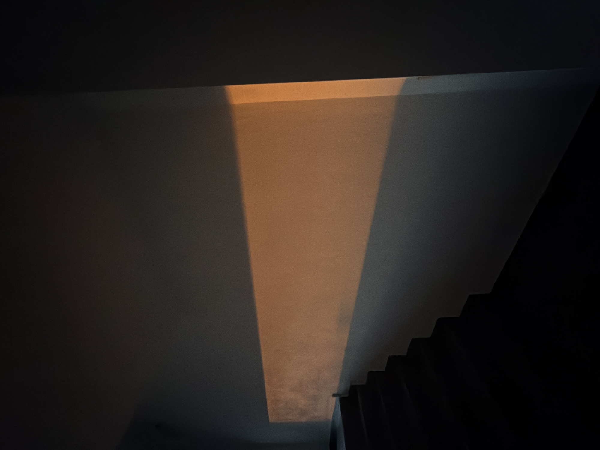 The image shows a stark, dimly lit interior with a long, horizontal band of warm, orange light illuminating a section of a plain, light-colored wall. To the left, a dark, stepped staircase recedes into shadow. The lighting suggests the sun is low in the sky, casting a dramatic shadow across the scene. The scene is devoid of any people or animals. The architectural style appears modern and minimalist. The specific location in the city of Nitra, Slovakia, is not identifiable from the image alone.
