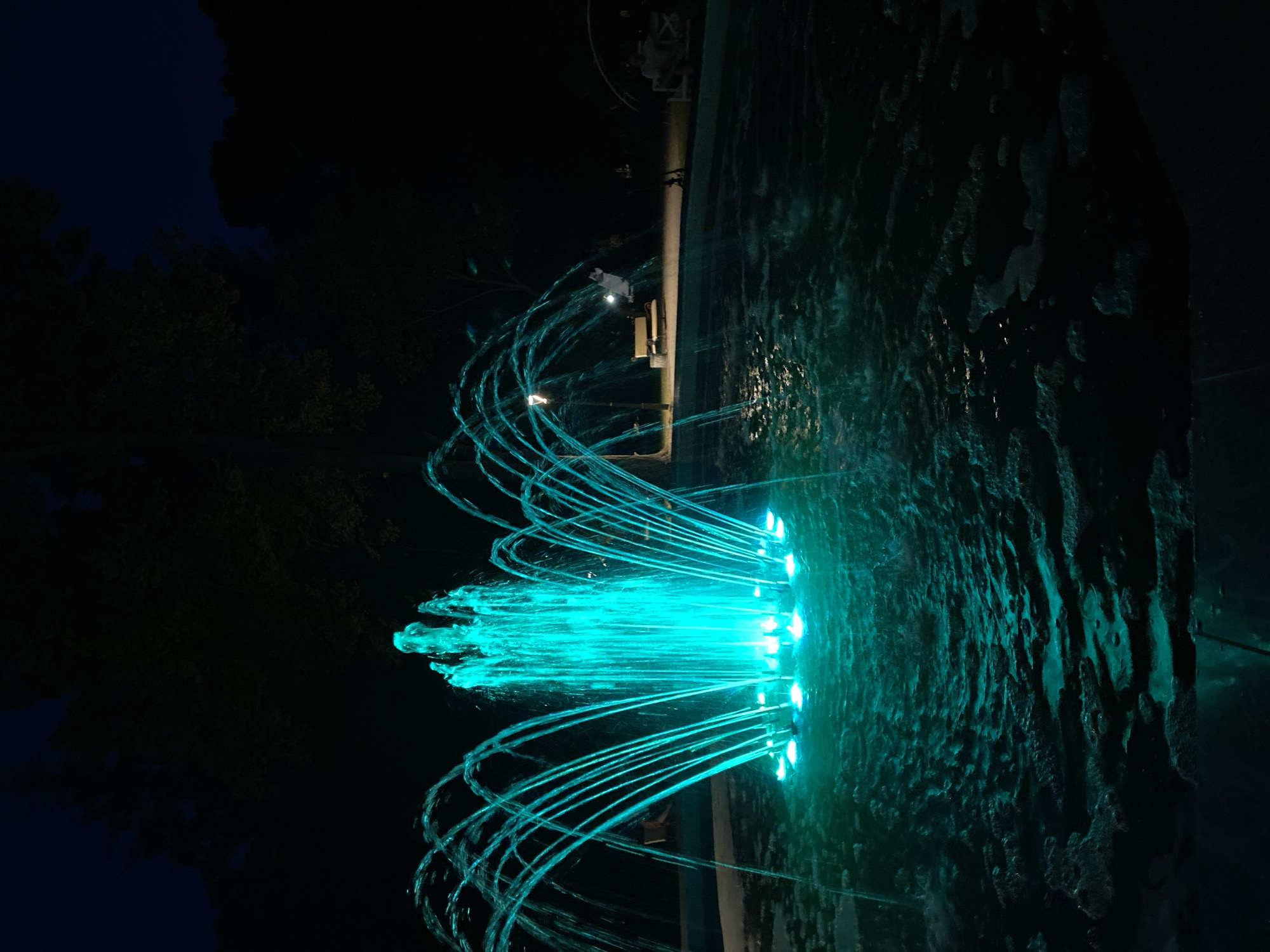 This is a nighttime image of a fountain with turquoise lighting. The fountain is in the foreground, with water shooting upwards and outwards in arcs. The water is illuminated from below, creating a glowing effect. In the background, there are trees and a dark sky. There are also a few figures visible in the background, sitting on benches and appearing to be enjoying the fountain. The scene is set in a park or public space. The location is the Feskovsky Square in Poltava, Ukraine, as indicated by the specific design and lighting of the fountain.