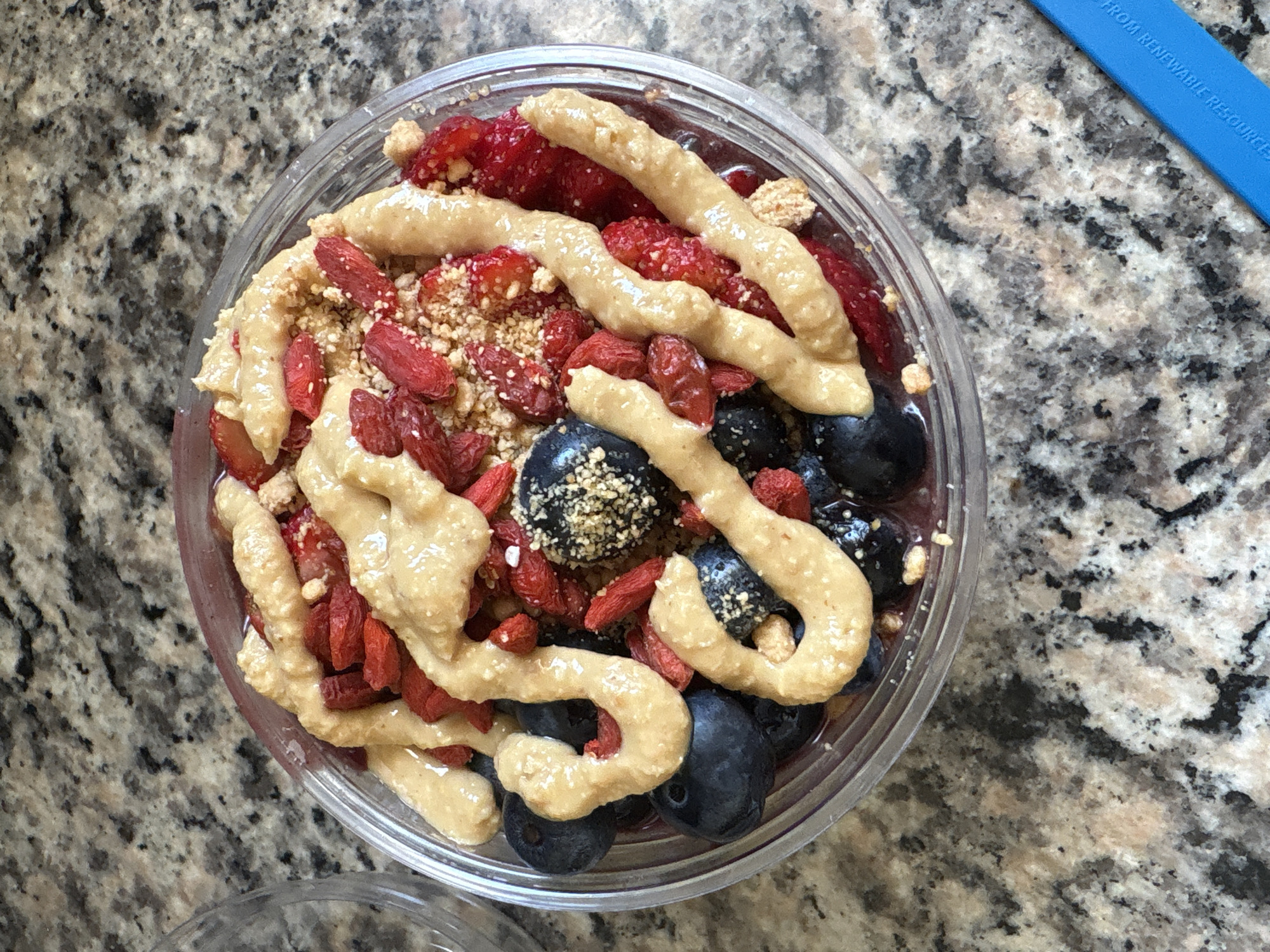A high-angle, close-up shot depicts a round, clear plastic bowl filled with an acai bowl, presumably in Seal Beach, California, United States. The bowl contains blueberries, chopped strawberries, goji berries, granola, and a light-colored, creamy topping drizzled in an irregular pattern. The bowl sits on a countertop featuring a speckled, beige-and-gray pattern. A blue, rectangular object with text partially visible ("FROM RENEWABLE RESOURCE...") appears in the upper right corner of the frame.