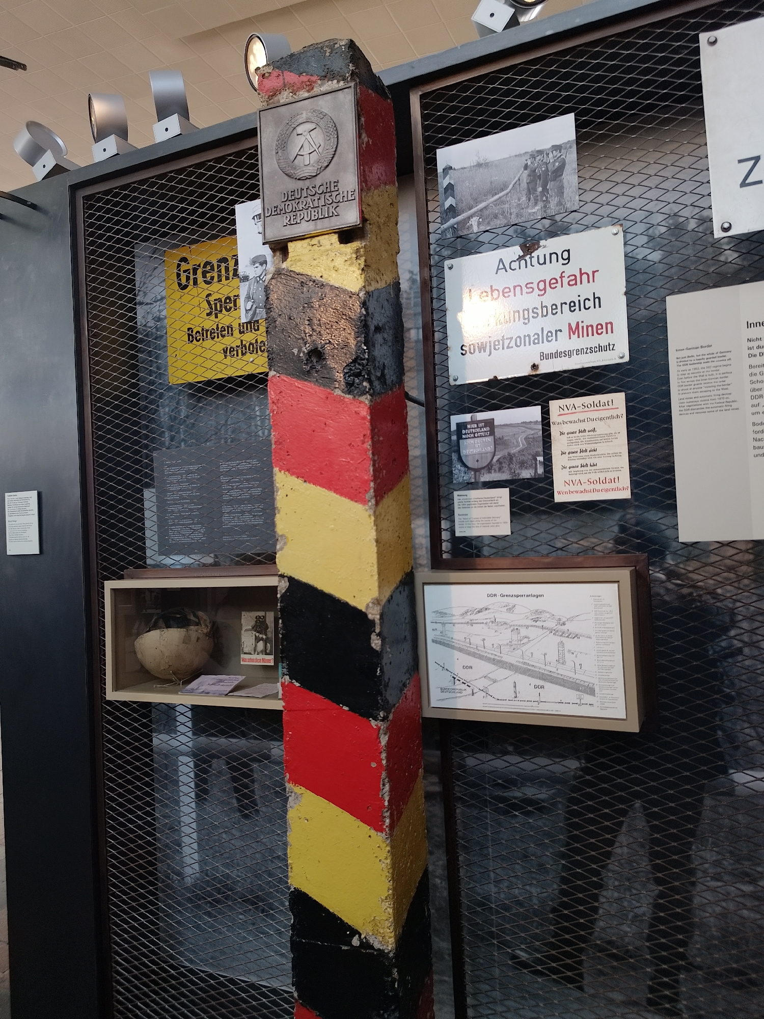 The image shows a display about the former German Democratic Republic (GDR) and the border between East and West Germany. The display features a concrete border marker painted with the colors of the German flag (black, red, and gold). On top of the marker is a plaque with the coat of arms and name of the GDR. Behind the border marker is a metal mesh wall with various signs and exhibits related to the border. These include a sign warning of landmines, photographs of border guards, diagrams of border fortifications, and text panels with information about the border and its impact. One of the exhibits contains a helmet and other artifacts. The overall impression is of a historical exhibit documenting the division of Germany during the Cold War.