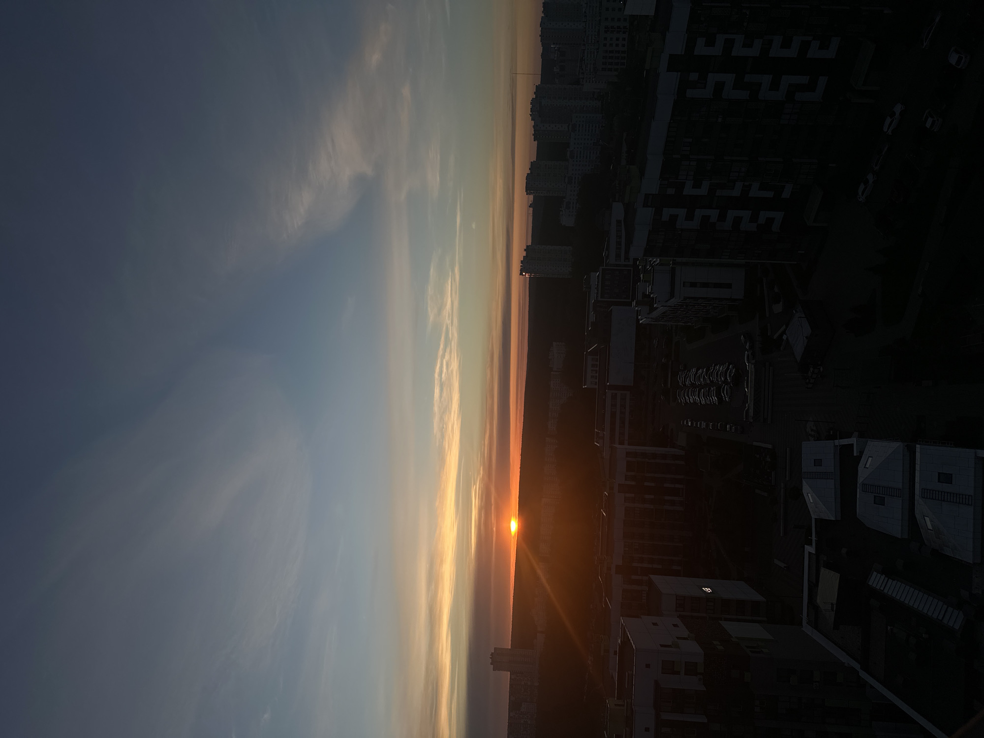 A panoramic view of a city at sunset or sunrise, potentially within Бараўлянскі сельскі Савет, Belarus. The sky exhibits a gradient from a pale blue to oranges and yellows where the sun is setting or rising. Buildings of various heights fill the right side of the frame. There is evidence of parking lots with cars within them among the buildings. The lighting is dark on the building side, suggesting the sun is behind them.