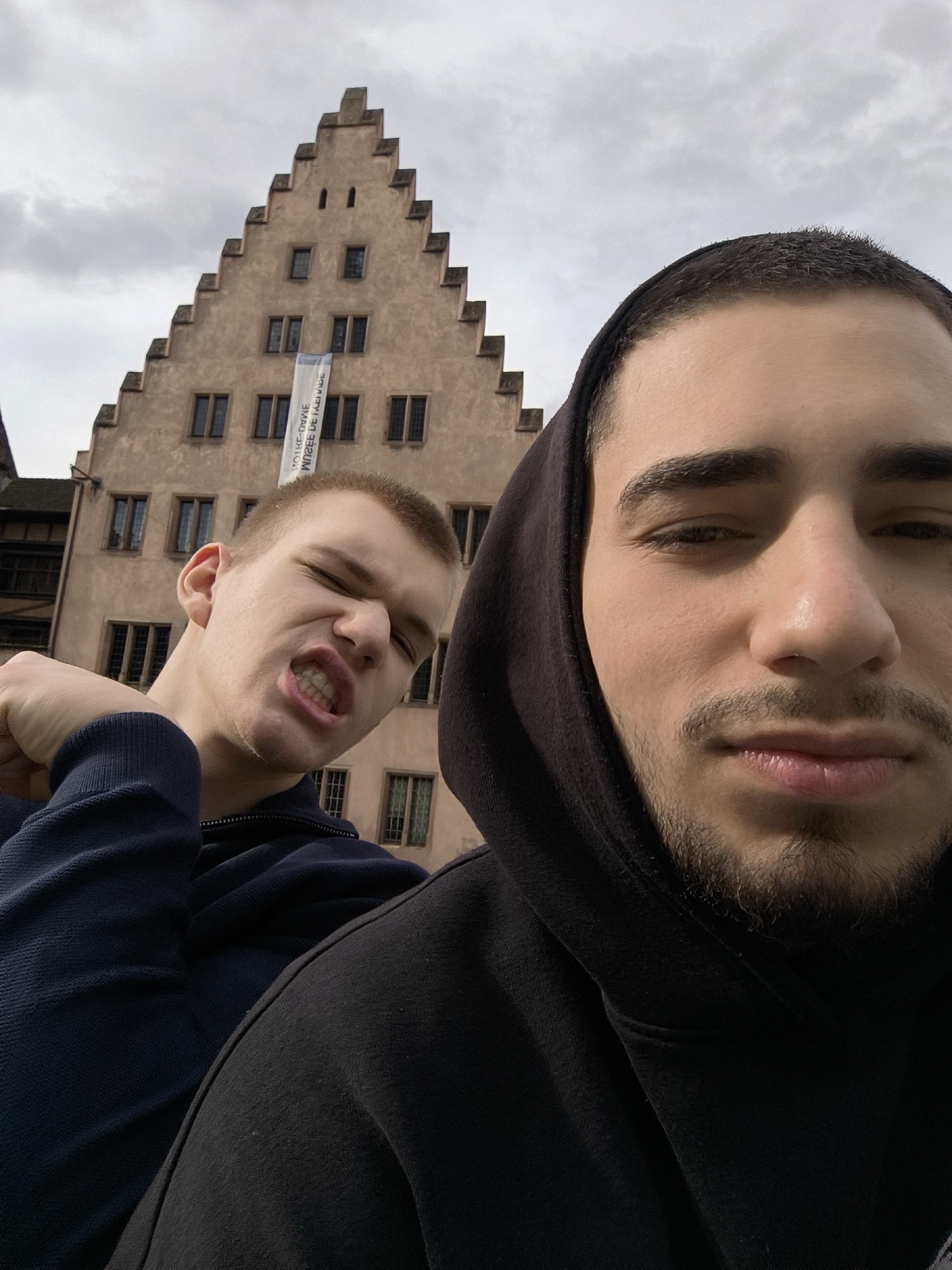 Two young men pose for a selfie in Strasbourg, France. The man on the right is wearing a black hooded sweatshirt, while the man on the left is wearing a dark blue top. Behind them is a multi-story building with a peaked roof, identified as the Musée de l'Œuvre Notre-Dame, displaying a banner vertically. The sky above is overcast.