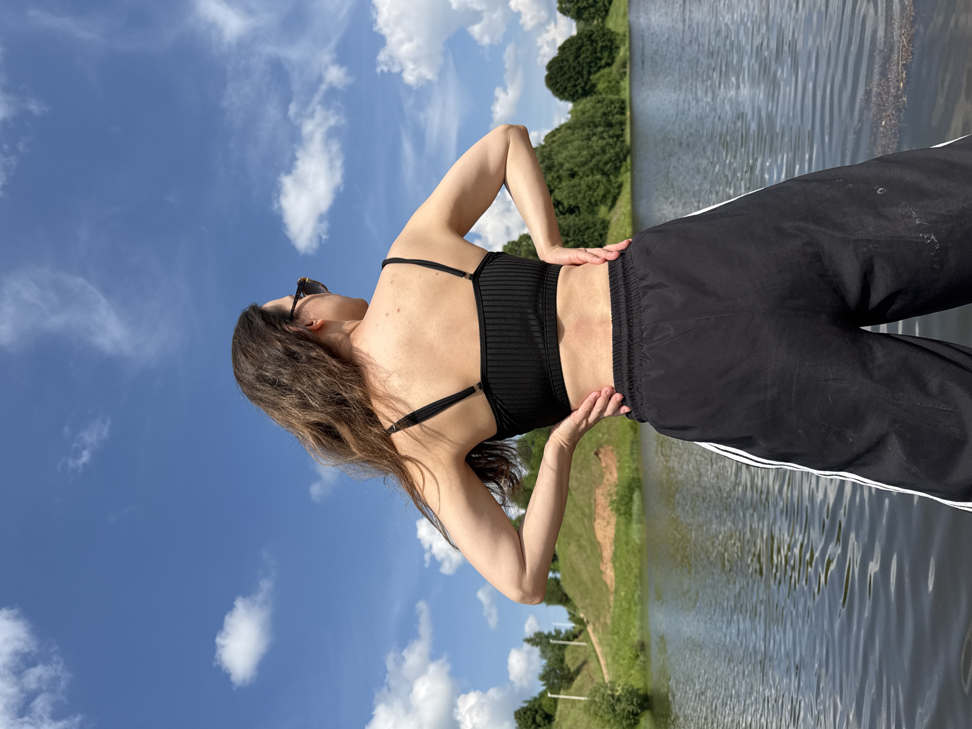 A full, distant rear view shows a person standing with their hands on their hips in front of a body of water, with a grassy, tree-lined hill and blue sky with clouds behind them. The person is wearing a black crop top and black athletic pants with white stripes down the sides. They are facing away from the camera, looking to the right. The environment appears to be a park or recreational area, possibly located in Zasechnyi, Russia, given the visual cues of a man-made incline with a walking path, common in some park designs.