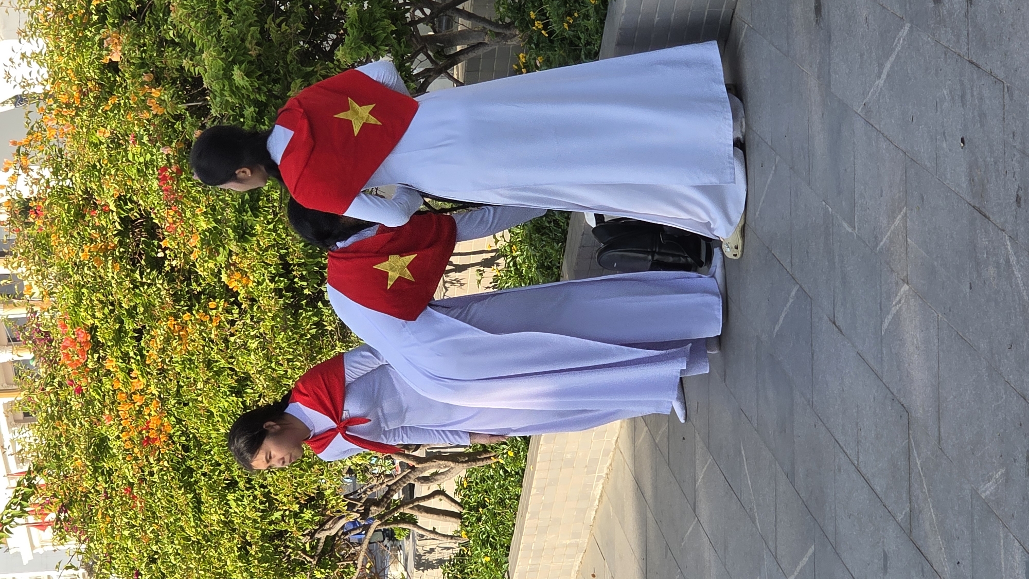 Three women in white áo dài dresses are standing outside on a paved surface. They are wearing red scarves around their necks that display a gold star. A bush with orange and yellow flowers is behind them. There is a building in the distance. The location is likely Nha Trang, Vietnam due to the dresses and the flags.