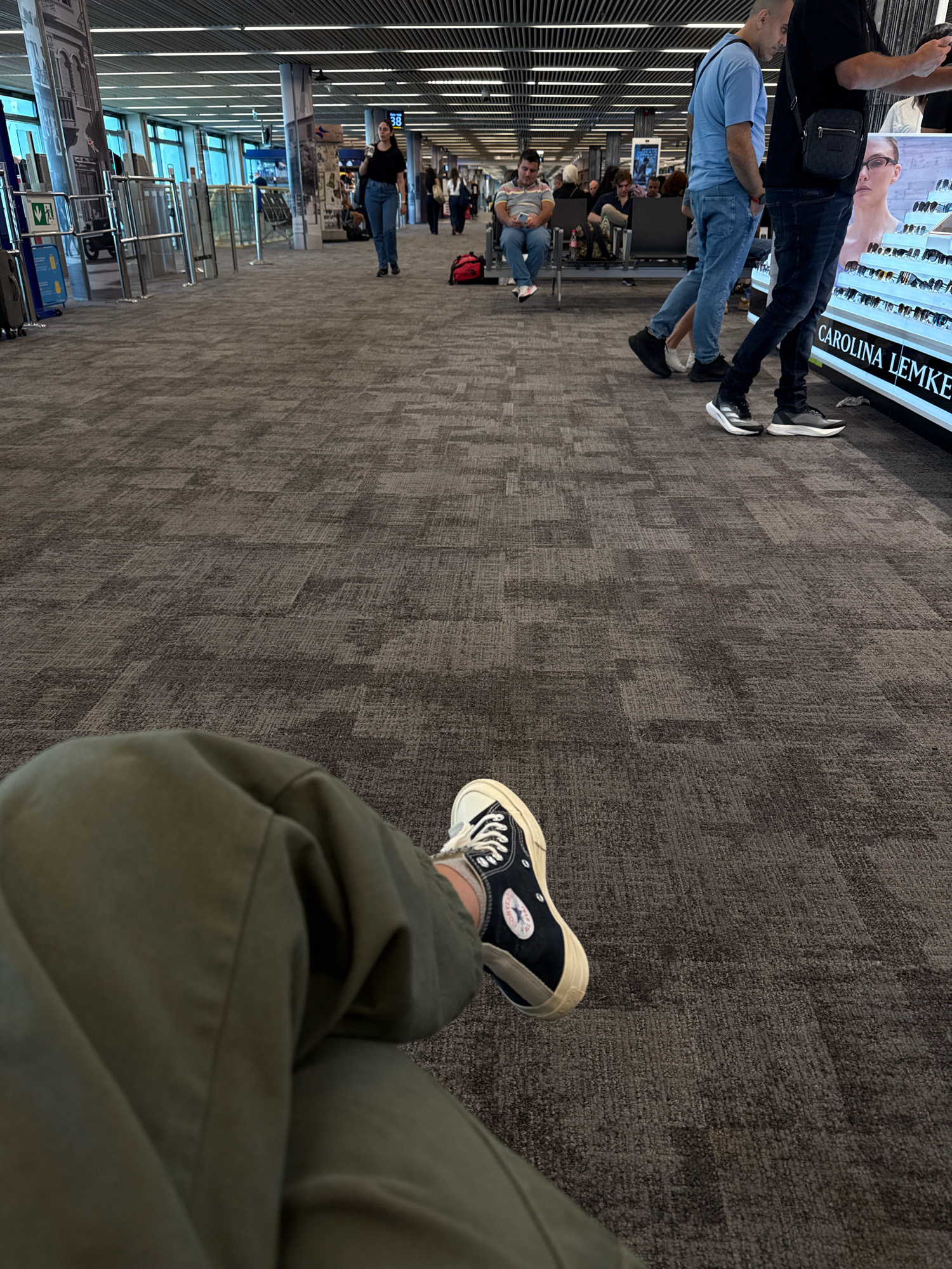 The image captures an indoor scene, likely an airport terminal, as indicated by the presence of seating areas, display cases, and what appears to be a gate or boarding area. In the foreground, a person's legs and feet are visible, wearing olive green pants and black high-top sneakers. The person is seated, and their gaze is directed downwards.
In the mid-ground, several other individuals are present. Two men are walking towards the right, one in a blue t-shirt and jeans, the other in a black t-shirt and dark jeans, carrying a messenger bag. Another man sits on a bench to the left, looking at his phone. In the background, more people are seated and walking, suggesting a busy environment. A large display case for eyewear, branded "CAROLINA LEMKE," is positioned on the right side of the frame. The flooring is carpeted with a dark, patterned design. The overall lighting is diffused and consistent with an indoor public space.
There is no indication that this location is in the city of Yagel, Israel.