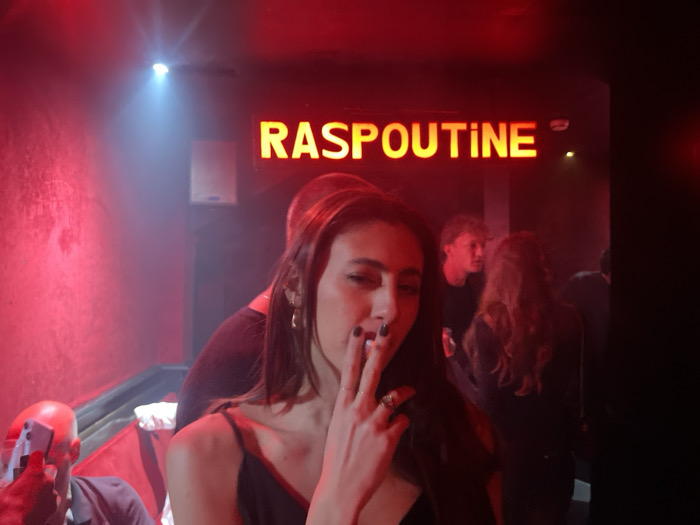 In a dimly lit interior with red lighting and some smoke, a woman with long, straight, dark hair holds a cigarette to her lips. The word "RASPOUTINE" shines brightly above her. In the background, several people are standing, including a man looking in her direction. Another man with a shaved head holds a phone up as though taking a picture. The overall mood is nightlife with a slightly edgy feel.