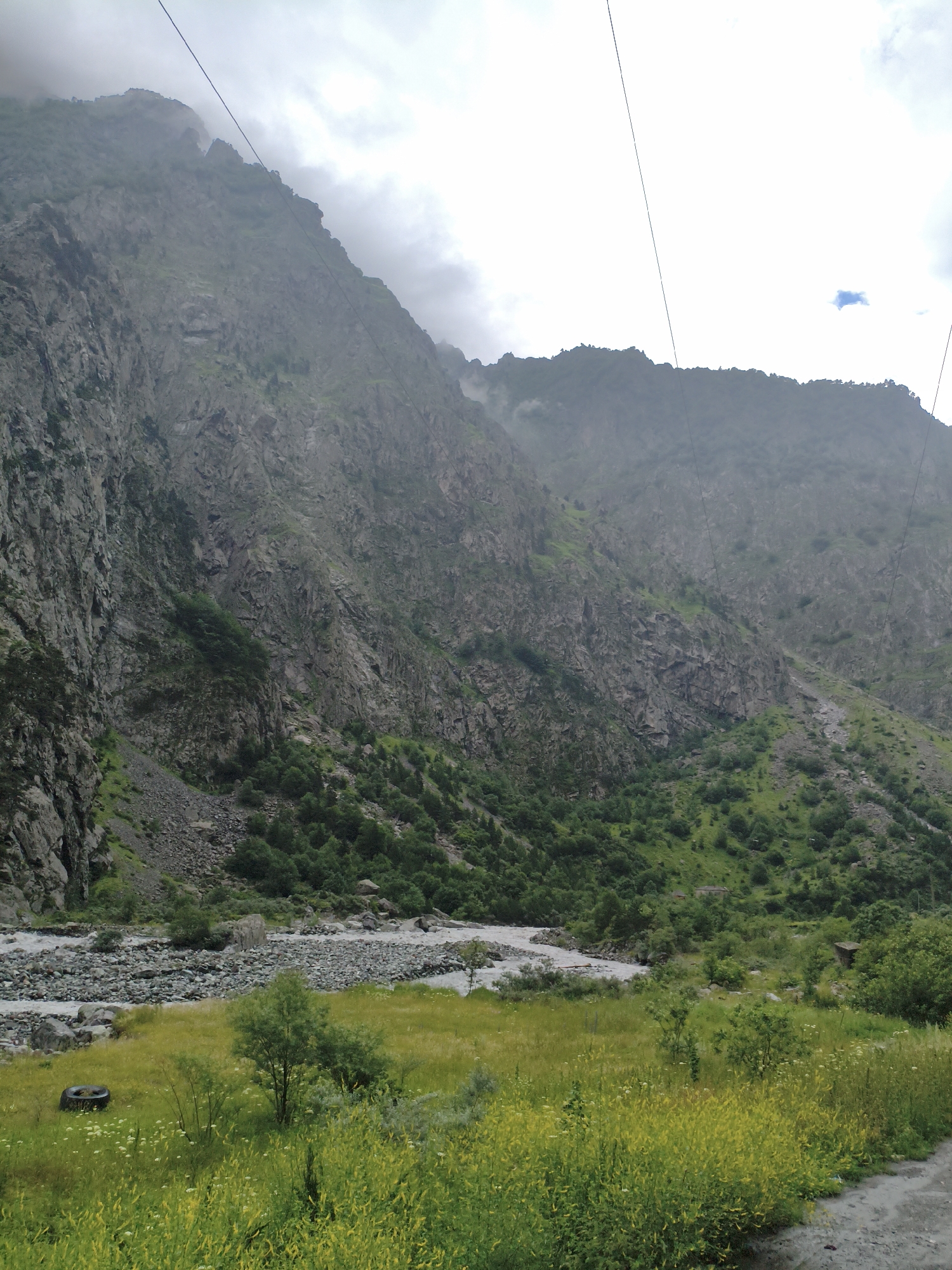 The image depicts a mountainous landscape, likely near Verkhnii Lars, Russia. A steep, rocky mountain face dominates the left side of the frame, with sparse green vegetation clinging to its lower slopes. A riverbed filled with stones runs through the valley floor, with a shallow, turbulent river flowing through it. In the foreground, a field of green grass and yellow wildflowers stretches across the scene, with a few small trees scattered throughout. A discarded tire is visible in the grass on the left. The sky is overcast with patches of light, suggesting a cloudy day. Power lines are strung across the upper portion of the image, suggesting human infrastructure in the otherwise natural environment. The overall impression is one of a remote and rugged natural setting.