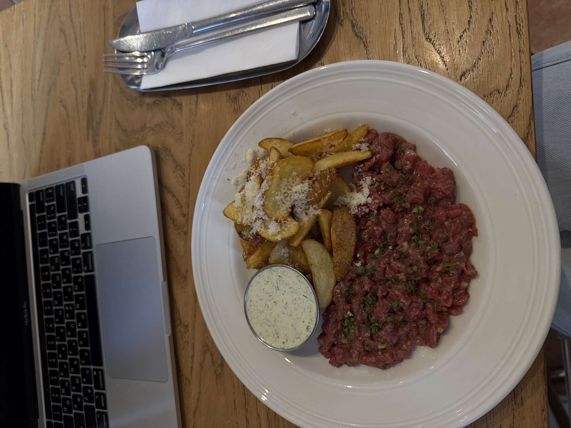 The image shows a plate of steak tartare with fries and a dipping sauce on a wooden table, next to a laptop and silverware. The steak tartare is arranged in a mound on the right side of the plate, sprinkled with green herbs. The fries are piled on the left side of the plate and topped with shredded cheese. The dipping sauce is in a small metal container. The laptop is a silver MacBook Pro, open and positioned on the left side of the frame. A set of silverware is arranged on a small silver platter with a white napkin, positioned in the upper left corner. The table has a light brown wood grain. The setting appears to be a restaurant or cafe. While precise coordinates are unidentifiable with the content of the image, it can be deduced that the image was taken in Belgrade, Serbia from the file's EXIF data (latitude: 44.8167, longitude: 20.4667).