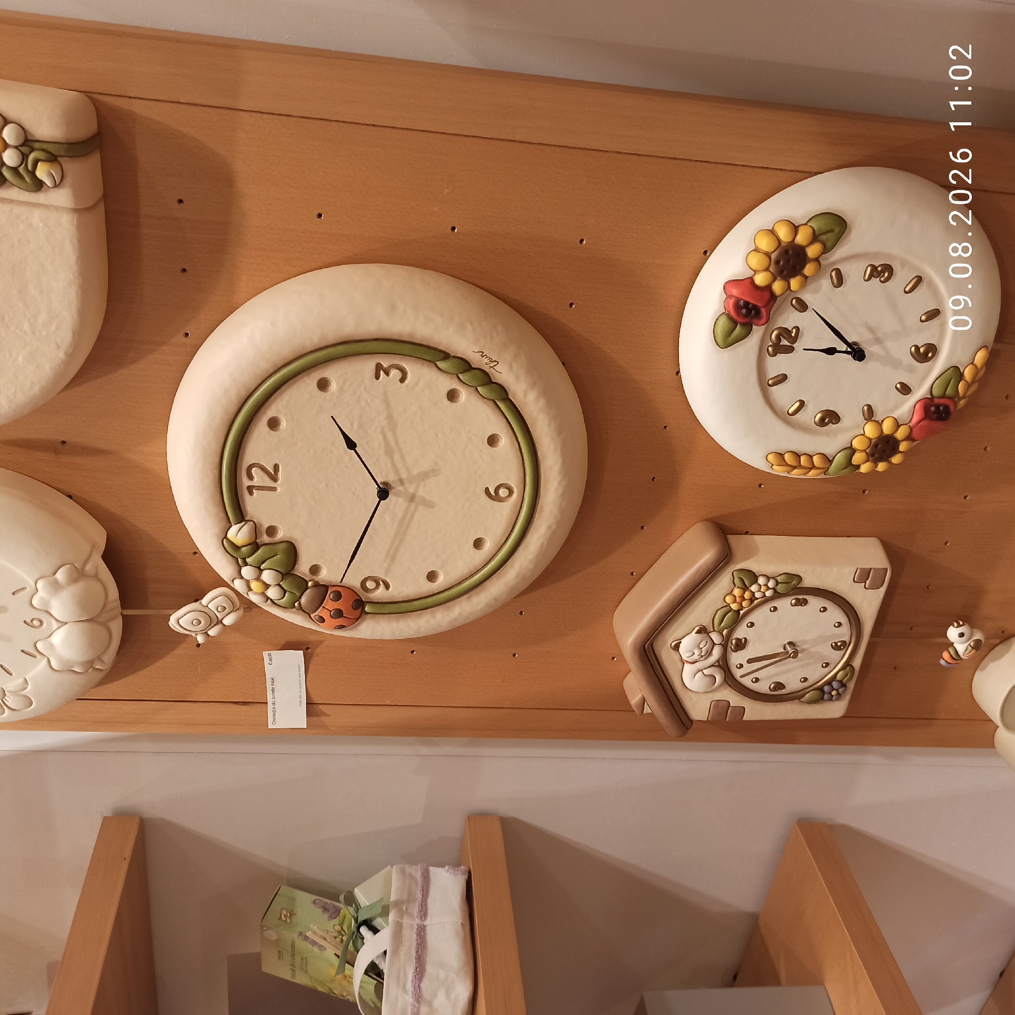 The image depicts a close-up, eye-level view of a wall decorated with ceramic clocks, likely in a retail setting. On the left, a wooden shelf holds a white basket with a light purple ribbon. To the right of the shelf, a large round clock with a light beige face and a green border is mounted on a wooden plank. This clock features a ladybug and flower embellishments. Below it, a smaller clock shaped like a house with a sleeping cat on the roof is visible. Further down, a small ceramic bird is hanging. To the right of the house-shaped clock is another large round clock with a similar beige face and floral decorations, including sunflowers and roses. The background consists of a light-colored wall and the wooden plank. The image is timestamped "09.08.2026 11:02". There are no people or discernible actions in the scene. Based on the provided information, this scene is located in Molfetta, Italy.