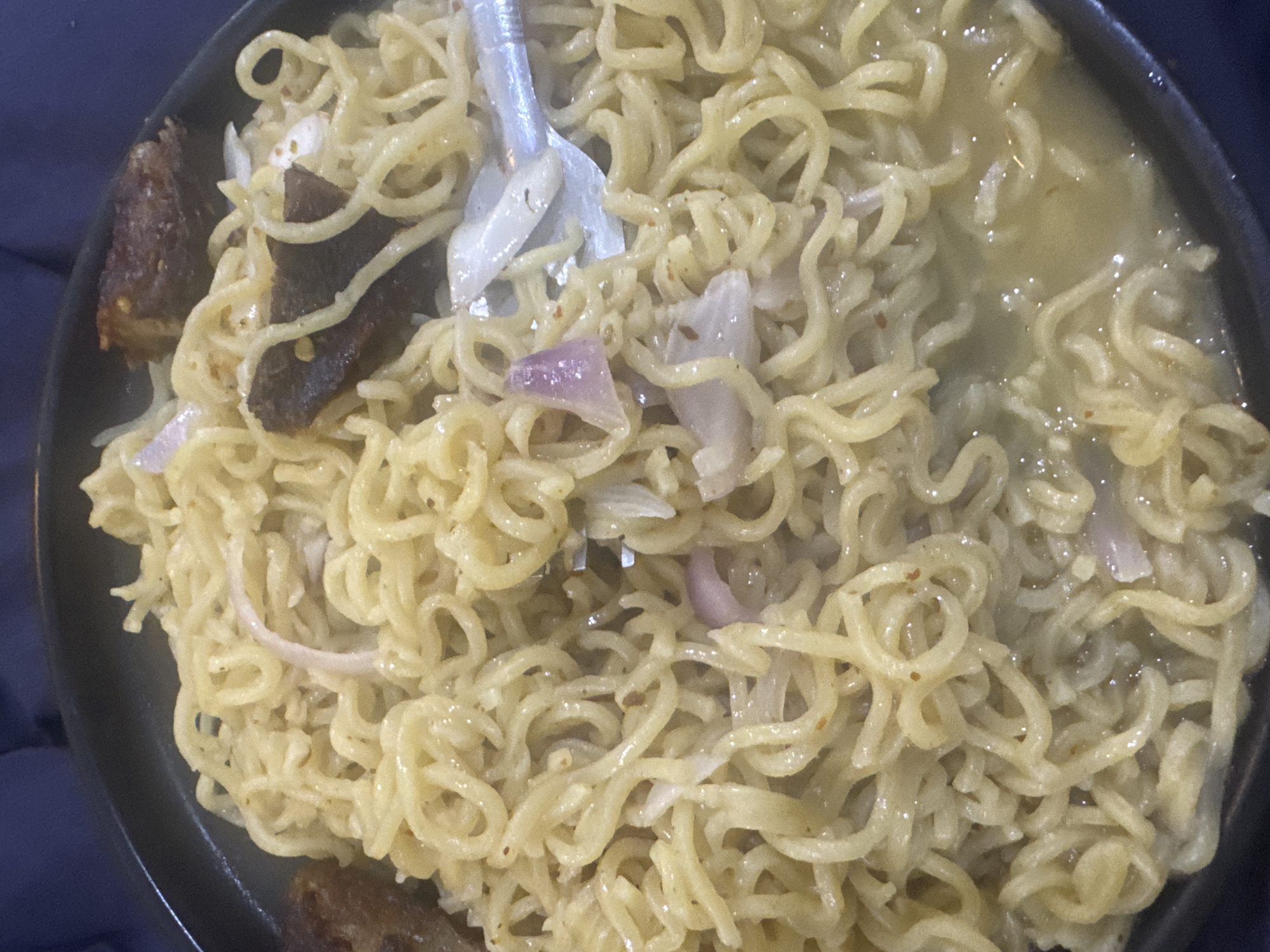 A close-up, overhead shot shows a black bowl filled with cooked instant noodles. The noodles are coiled and appear to be coated in a light, soupy sauce. Several pieces of dark brown meat, possibly beef or lamb, are mixed in with the noodles. Thinly sliced pieces of red onion are also visible, interspersed throughout the dish. A fork with a silver handle and tines is partially submerged in the noodles on the right side of the frame, suggesting the food is being eaten. The background is a dark blue fabric, possibly a tablecloth or clothing. The scene is indoors, and the lighting is consistent with artificial light. There is no visible indication of the specific location within Abuja, Nigeria, other than the food itself.