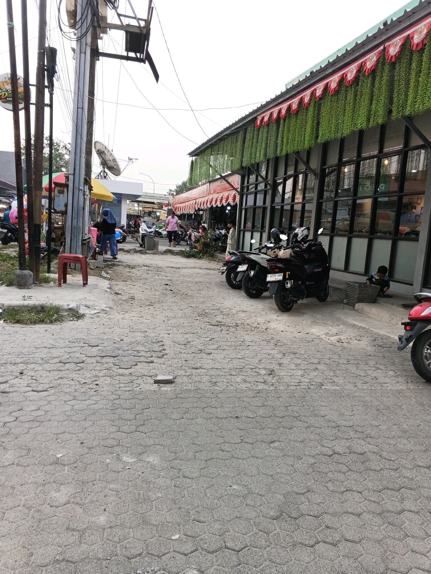 The image depicts a street scene in Cikande, Indonesia, on a cloudy day. The foreground is dominated by a paved street with a hexagonal block pattern, showing some signs of wear and tear with broken and uneven sections. To the left, a utilitarian metal pole structure supports numerous electrical wires and a satellite dish. A small red plastic stool sits on the pavement near the base of the poles. Further left, a section of a market or roadside stall is visible, with colorful balloons and umbrellas.
In the middle ground, a paved walkway leads towards a row of buildings with dark, reflective windows. Several motorcycles are parked along the walkway. One person, wearing a blue headscarf and dark clothing, stands near a white structure with a yellow awning. Another person, in a pink shirt, walks away from the camera towards the buildings. The buildings themselves have a tiled roof adorned with red and white pennants and an abundance of hanging green artificial foliage, suggesting a restaurant or commercial establishment.
In the background, more parked motorcycles and people are visible, indicating a busy area. A young child is seated on the ground to the right of the buildings, near a patterned concrete planter. A red motorcycle is partially visible on the far right edge of the frame. The overall impression is a lively commercial street with a mix of traditional market elements and modern storefronts.