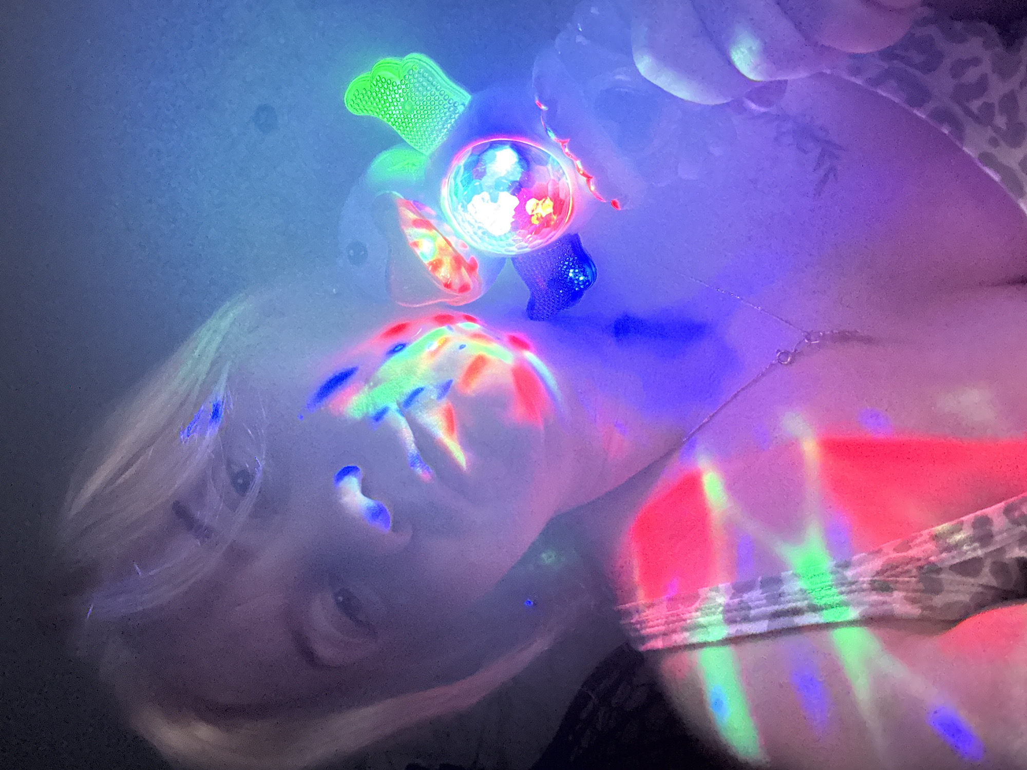 The image shows a person with light hair, possibly female, holding a small, lit disco ball in their hand. The disco ball casts colorful light patterns across the person's face, neck, and shoulder. The person is wearing a patterned top. A thin necklace is visible around the person's neck, and a tattoo is also visible on the person's shoulder. The overall lighting is dark except for the colorful reflections from the disco ball. The location in Chaikovsky, Russia, is not identifiable from the content of the image.