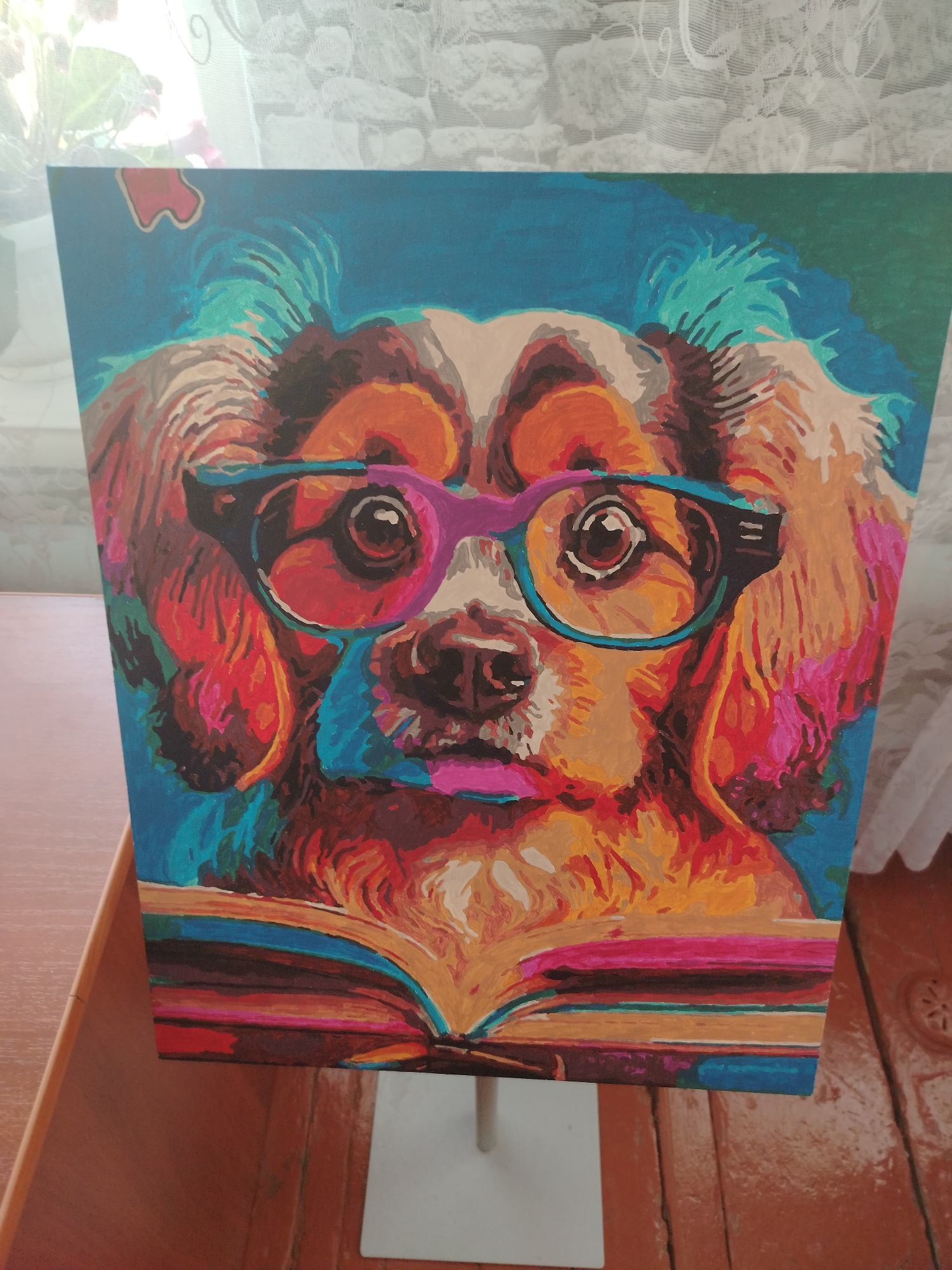 A vibrantly painted canvas depicts a dog wearing glasses and reading a book. The artwork is displayed on a white easel, positioned on a wooden surface. The background consists of a window with white lace curtains. The scene appears to be indoors.