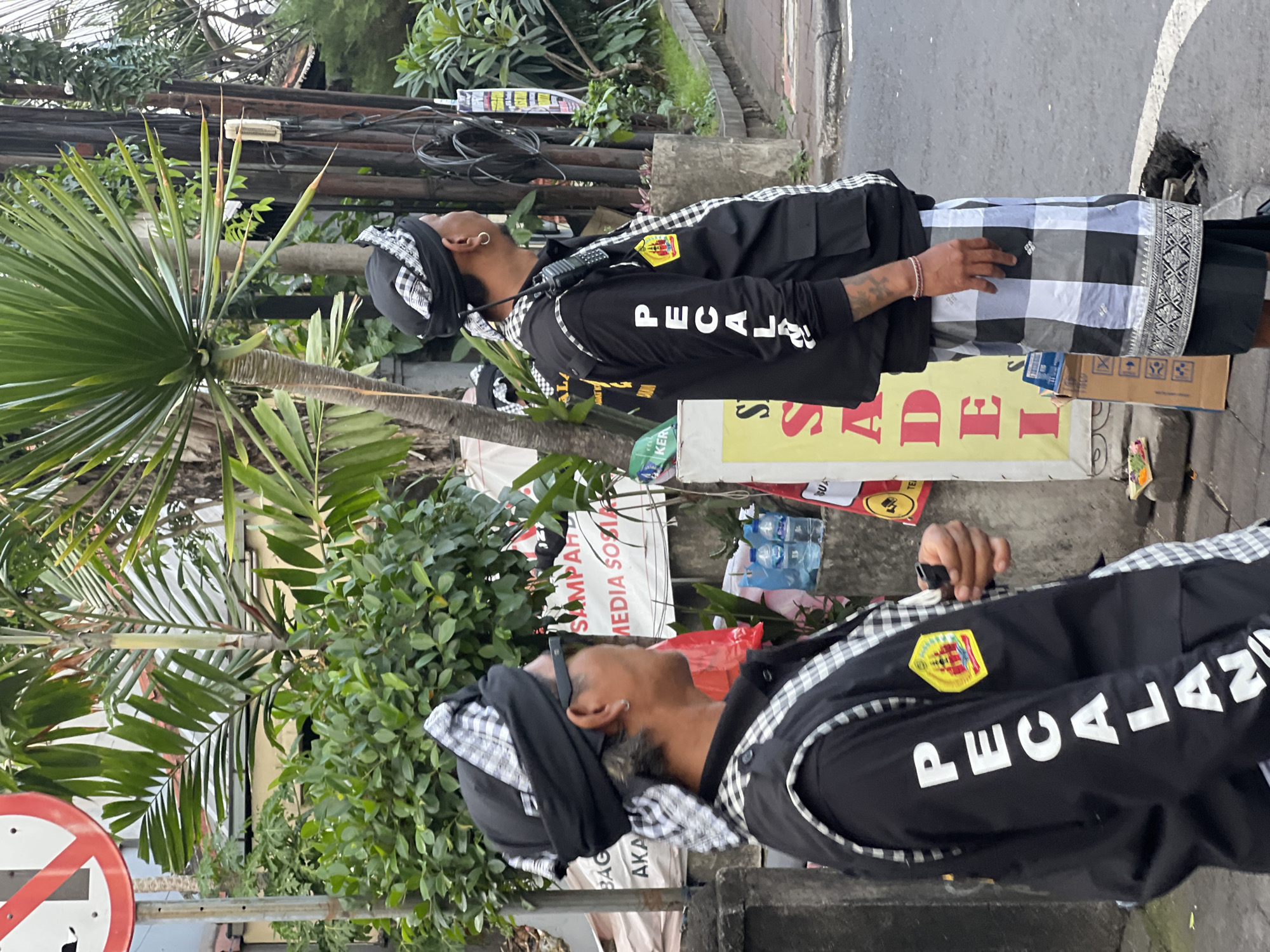 In North Kuta, Indonesia, two men stand on a paved street, possibly a sidewalk, near a building with a faded sign. The man on the left, facing right, wears a black vest over a black shirt. His vest has "PECALAN" printed vertically in white on the left sleeve and a yellow emblem with red and black imagery on the left chest. He also wears a black headwrap with a white and black checkered pattern and sunglasses. The man on the right, facing left, wears a similar black vest over a black shirt with the word "PECALAN" printed vertically in white on his left sleeve. He has a black headwrap with a white and black checkered pattern, a silver hoop earring, and a radio attached to his vest. He is holding a black object in his right hand. Both men are wearing traditional checkered sarongs. Behind them, there are green plants, including palm trees, and a red and white street sign. A yellow sign with red lettering is partially visible. Several bottles of water and other items are stacked on a concrete ledge. The overall scene suggests an outdoor gathering or event in a tropical urban environment.