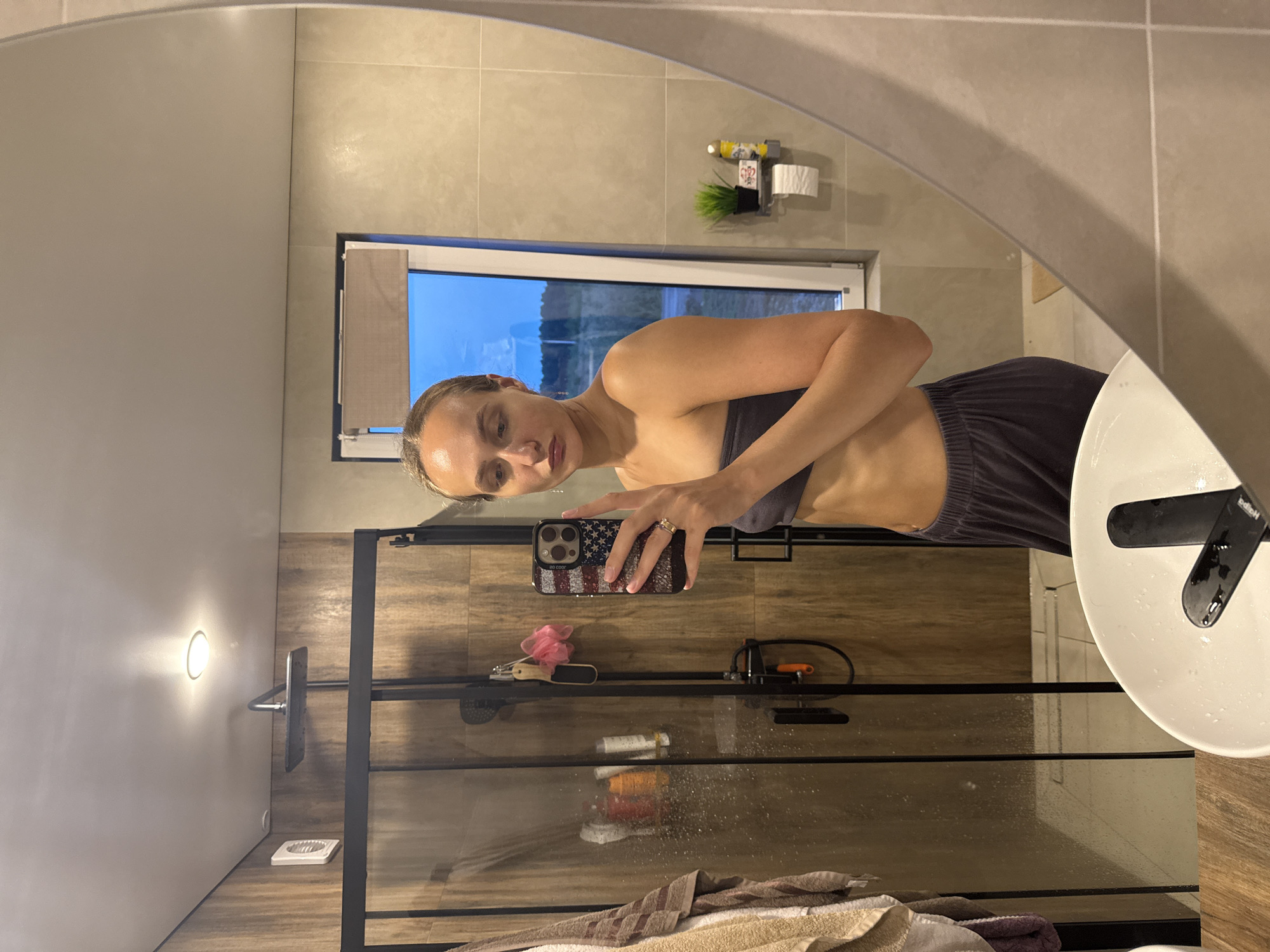 A woman is taking a mirror selfie in a modern bathroom. She is wearing a grey strapless top and dark grey lounge pants. Her blonde hair is pulled back, and she is holding a phone with an American flag-themed case. In the background, a glass-enclosed shower with wooden-tiled walls is visible. A sink is in the foreground, and a window showing a twilight sky and rural landscape is to the right. The bathroom is well-lit, with a ceiling light illuminating the shower area. The overall impression is of a private, clean, and contemporary space.