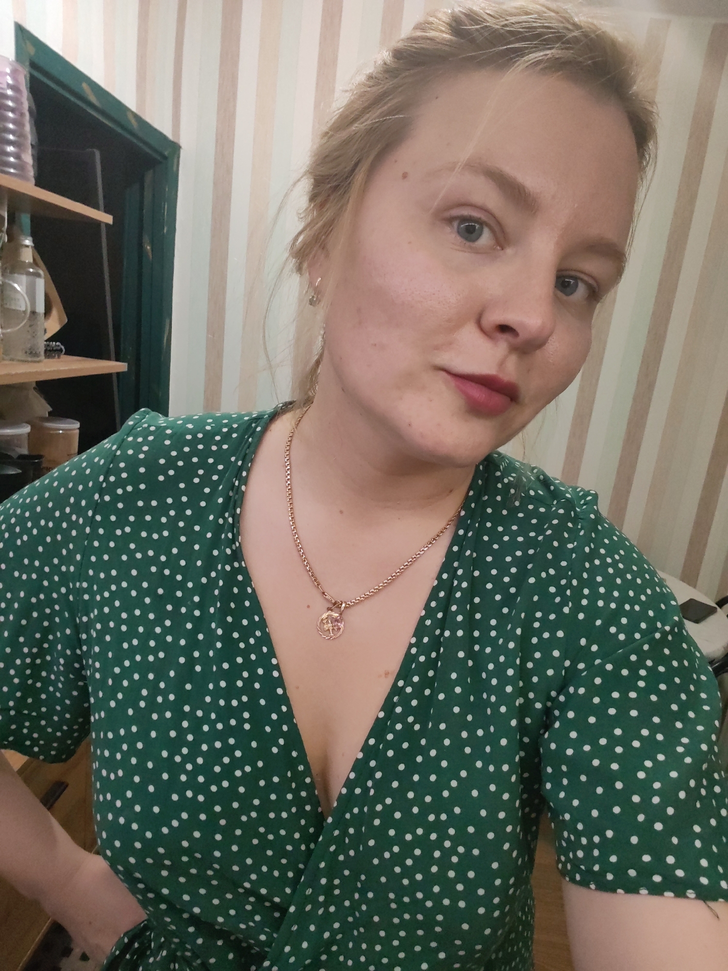 A woman is posing for a selfie indoors. She has fair skin, light blonde hair styled loosely, and blue eyes. She is wearing a green dress with white polka dots and a delicate gold necklace with a circular pendant. The dress has a V-neck. She is looking directly at the camera with a slight smile. The background consists of vertically striped wallpaper, part of a dark-colored doorframe, and a shelf with various objects, including glass bottles and jars.
There is no contextual information available in this image to indicate its location in the city of Izhevsk, Russia or any other specific location.