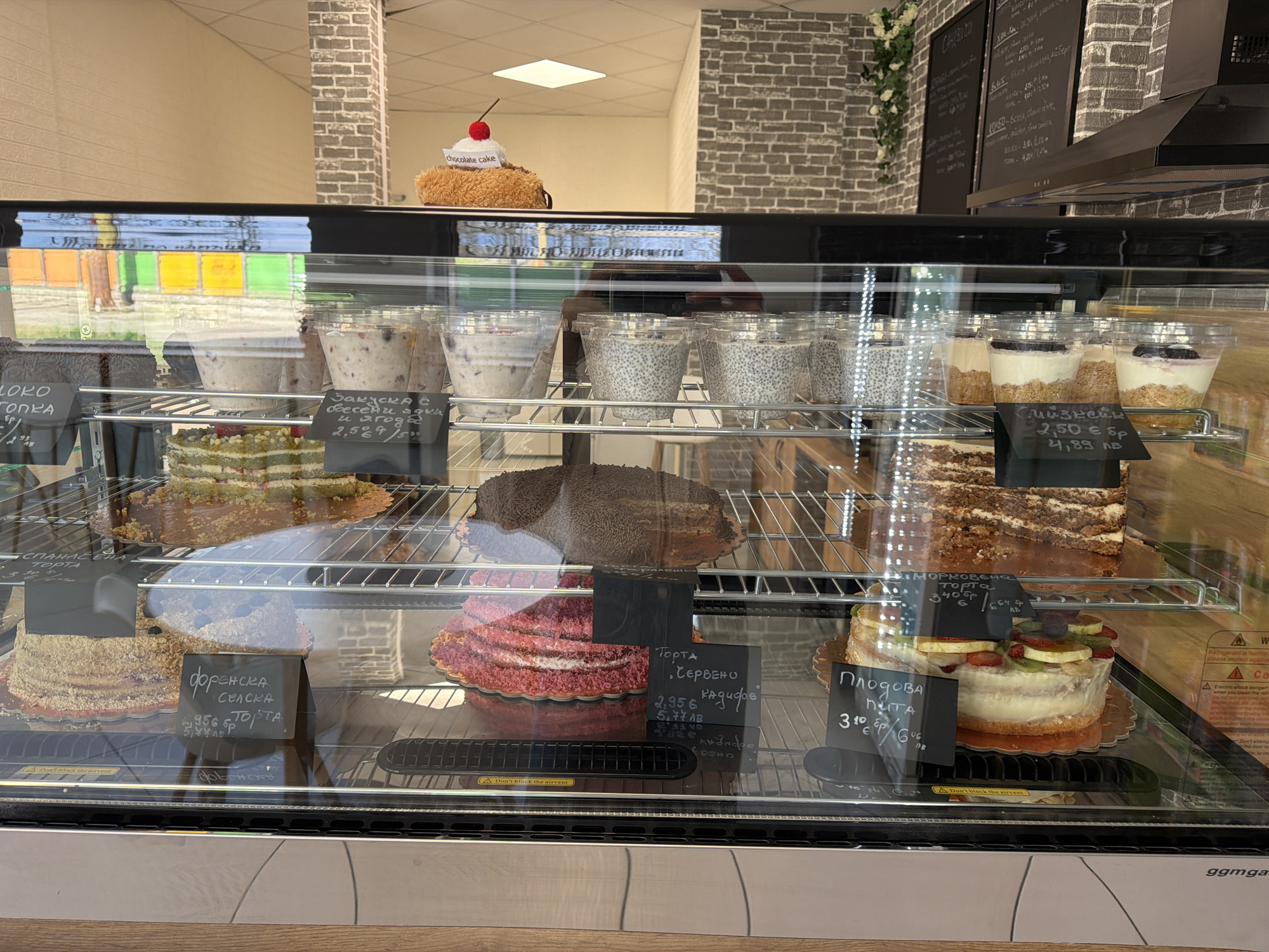 The image shows a refrigerated display case filled with various cakes and desserts. The scene appears to be inside a bakery or cafe, likely in Gabrovo, Bulgaria based on the Cyrillic text on the price tags and signs.
The display case has two shelves. The top shelf contains cups filled with chia seed pudding and other parfaits. The lower shelf holds several cakes, including a red velvet cake, a "forest" cake, a fruit tart, and other varieties with chocolate and nut toppings. Each cake has a small black sign in front of it, displaying the name of the cake and its price in both Euros and Bulgarian Lev.
Above the display case, there's a shelf with a stuffed animal that has a sign on it that says "chocolate cake". Behind the shelf, a brick wall is visible. Further up, a menu board with chalked text can be seen. The light reflecting off the glass of the display case makes it difficult to see all the details clearly.