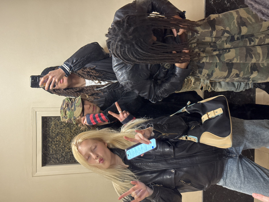 The image captures four individuals in an indoor setting, characterized by plain, light-colored walls and a dark, polished floor. A rectangular, dark, textured panel is recessed into the wall in the background.
On the far left, a blonde female with long hair wears a black leather jacket and light-wash jeans. She holds a light blue smartphone vertically in her right hand, its screen displaying content, and forms "rock on" gestures with both hands. Her eyes are closed, and her lips are pursed. A black and beige shoulder bag with light blue accents hangs from her left shoulder.
Behind her, a male wearing a camouflage-patterned cap and a dark jacket with a red and blue striped wristband on his left arm forms a peace sign with his left hand.
To the right, a third individual with long dark dreadlocks wears a dark jacket with white stripes on the sleeves over a white t-shirt and a necklace. This individual holds a dark smartphone up, obscuring their face, indicating they are taking a mirror selfie.
In the foreground on the right, a fourth individual, also with long dark dreadlocks, wears a black quilted leather jacket and camouflage-patterned wide-leg pants. This person is looking downwards, appearing to adjust their clothing.
The scene provides no identifiable landmarks or features to determine the location within Paris, France.