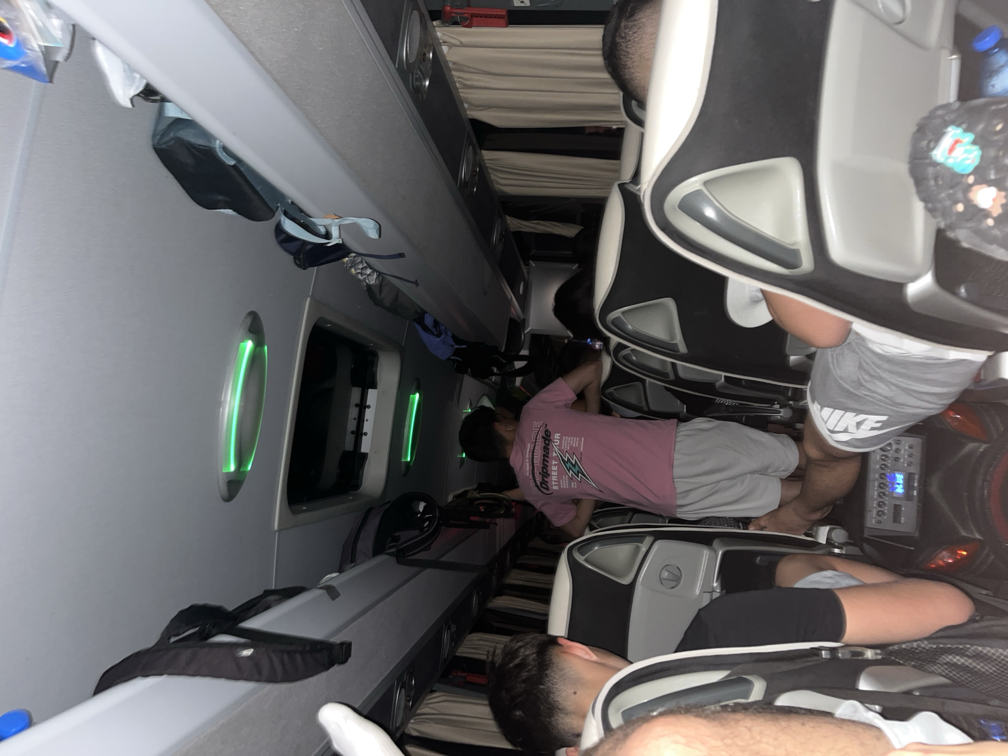 The image shows the interior of a sleeper bus. Passengers are seated in reclining seats with luggage in the overhead compartments. One person wearing a pink shirt stands in the aisle. Another person in grey shorts and a "NIKE" shirt stands near a control panel. Other passengers are reclining in their seats. The exact location within Dinkata, Bulgaria, is not discernible from the image.