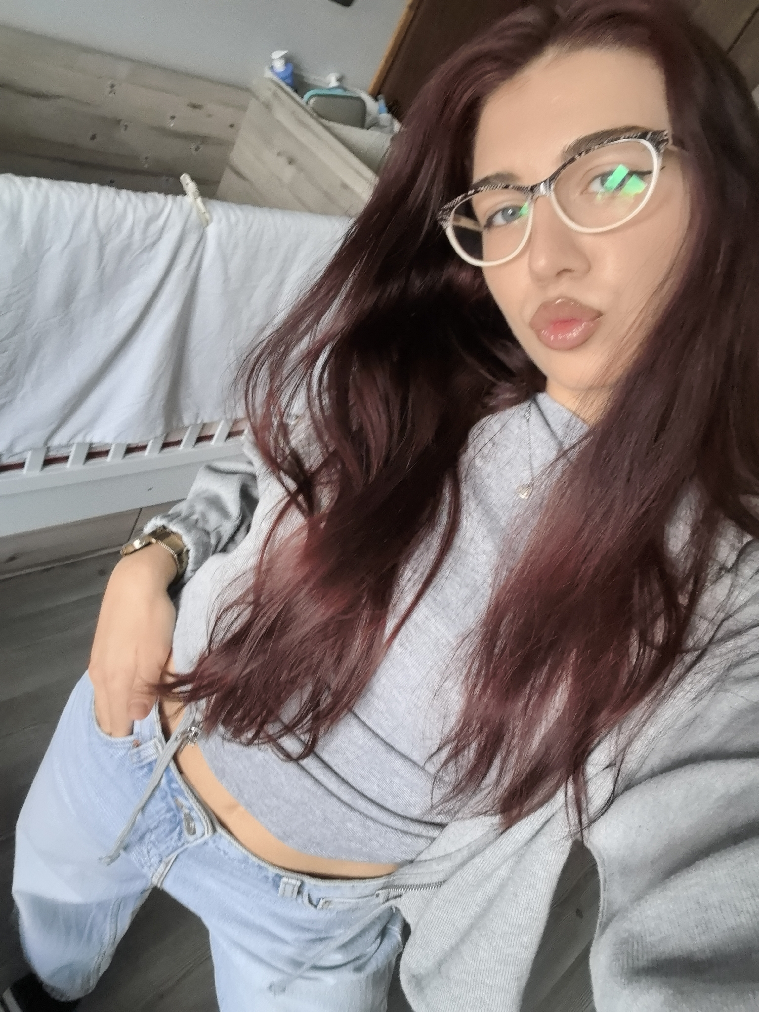 A young woman with long, dark reddish-brown hair is captured in a selfie, posing indoors in what appears to be a residential setting in Stara Zagora, Bulgaria. She wears white and black patterned eyeglasses that reflect a green light source. Her lips are glossy and slightly pouted, and her gaze is directed towards the camera.
She is dressed in a light grey, long-sleeved crop top that reveals a sliver of her midriff, paired with light blue denim jeans that are unbuttoned and partially unzipped on one side. One hand is tucked into her jeans pocket, while a gold-toned watch with a dark strap is visible on her other wrist. A delicate necklace with a small pendant can be seen around her neck.
The background includes a white sheet or fabric draped over a bed-like structure with a white wooden frame. A single clothes peg rests on the sheet. Behind this, wooden wall paneling is visible, and on a shelf or surface in the upper background, there are a few bottles or containers. The floor appears to be wooden. The lighting suggests it is daytime. No text is visible in the image.