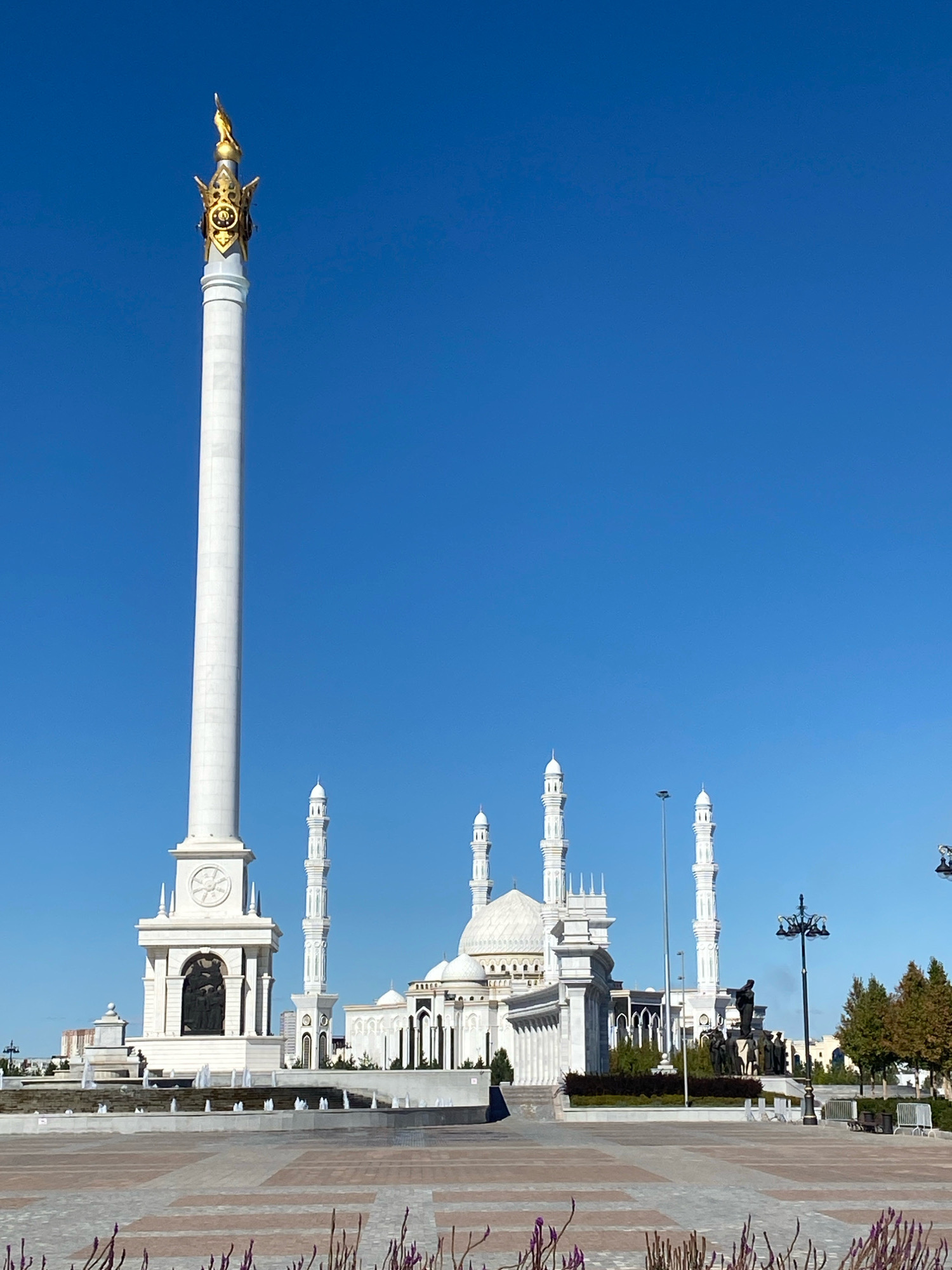 The image depicts a wide shot of the Bayterek Monument and the Hazrat Sultan Mosque in Astana, Kazakhstan, under a clear, bright blue sky. The Bayterek Monument, a towering white structure with a golden orb at its apex, dominates the left side of the frame. Its base features ornate carvings and a circular design. In the mid-ground, the white Hazrat Sultan Mosque stands with its multiple domes and minarets. The mosque is flanked by lush greenery and other architectural elements. In the foreground, a paved plaza with intricate patterns stretches out, with some sparse, dry foliage visible at the very bottom. Several lampposts and a distant statue are also present in the scene. There are no people visible in the image. The overall impression is one of grandeur and architectural prominence.
