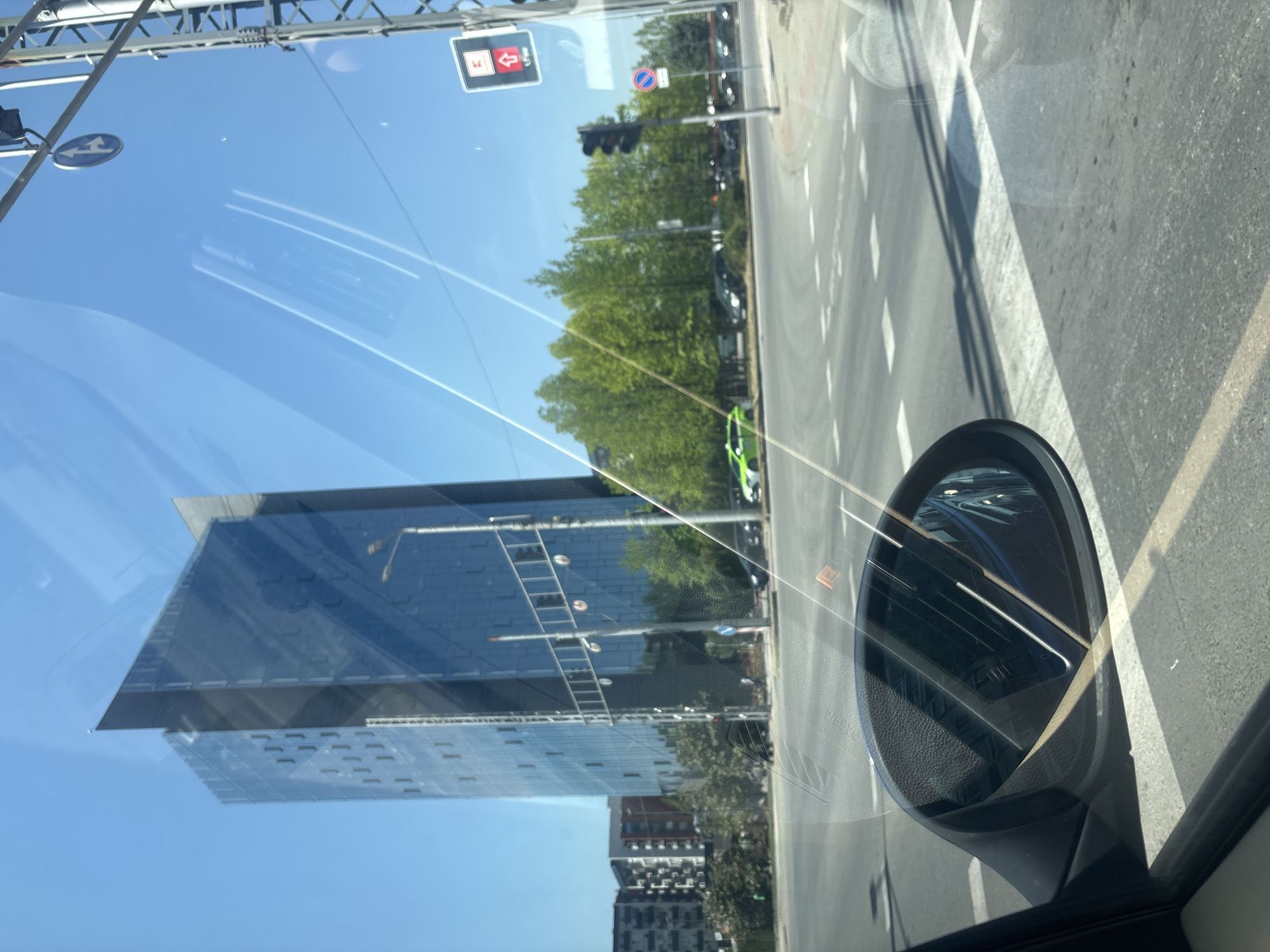 The image captures an urban street scene from the perspective of a vehicle's interior, likely through the windshield. The overall setting is a bright, sunny day in what appears to be a modern city environment.
In the foreground, on the right side, the black casing and glass of a car's side-view mirror are visible. Reflections of the surrounding environment, including what looks like a blue vehicle, are seen within the mirror. The windshield itself shows various reflections, creating a layered effect over the exterior scene.
Looking out, on the left side of the frame, stands a striking modern building with a facade composed largely of blue-tinted glass panels, reflecting the clear blue sky and some surrounding elements. Below this main structure, another building with multiple windows is partially visible.
The center and right side of the image show a tree-lined street or avenue. Lush green trees create a natural barrier along the road. Several cars are visible on the street, including a distinct bright green car parked or driving among the trees. The asphalt road features white dashed and solid lines.
Above the street, various traffic infrastructure elements are present. These include a black traffic light showing a red signal, an overhead gantry structure, and several road signs. One sign is a blue circle with a white left-turn arrow, another is a red circle with a horizontal white bar (no entry/forbidden), and a distinct red square sign with a white arrow pointing upwards and a smaller arrow to the right. The sky is a clear, vibrant blue, indicating good weather. Strong shadows from overhead structures are cast across the road surface, consistent with bright sunlight.
No people are distinctly visible. The time of day is clearly daytime, given the bright illumination and strong shadows. There is some text visible on the side-view mirror casing that is difficult to decipher, but parts resembling "YILPYC" might be present. The specific traffic signs offer potential location cues but are not definitive without further context.
