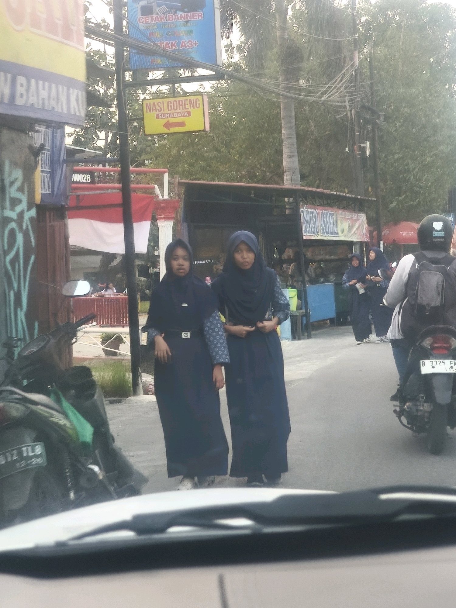 In Bekasi, Indonesia, two young women wearing blue hijabs and long dark dresses stand on a paved street. The woman on the left is facing forward and has her hands on her waist. The woman on the right is facing slightly away from the camera and is holding something in her hands. Behind them, a motorcycle is parked with a green bag on its handlebars. In the background, there are buildings, trees, and electrical wires. A red and white Indonesian flag is visible hanging from a pole. A yellow sign with red text reads "NASI GORENG SURABAYA" with an arrow pointing to the right. Another sign on a building reads "BAHAN KU" with some text below it. The scene appears to be during daylight hours, possibly late afternoon given the lighting.