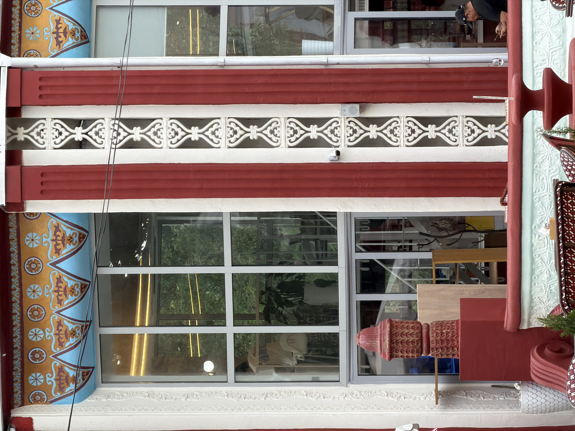 This image captures a brightly decorated building facade with an active outdoor seating area, located in Unknown, Unknown.
The building's facade is characterized by a vibrant color palette of red, white, blue, and gold. Prominent broad, fluted red pilasters alternate with narrower white sections. A central white vertical panel features an intricate, openwork, repeating pattern of white scrolled elements. A small white security camera and a compact white speaker are mounted on this central panel. Above the large, multi-paned windows, a decorative frieze displays a golden band with circular floral motifs in red and blue, topped by a dark red border. Below the golden band, a light blue section is adorned with symmetrical, stylized scrollwork in gold and red. The windows themselves are white-framed and reflect external greenery and internal ambient lighting, including linear yellow LED strips.
Through the left window, an indistinct person in a light-colored top is visible standing inside, along with a white bag and a large potted plant. Through the right window, a white ribbed vase or pot is discernible.
In the foreground, an elaborate outdoor seating area is defined by a low, ornate red barrier featuring a light blue-green section with white relief patterns. Centrally positioned on this barrier is a tiered, red pedestal-like structure, possibly a small fountain or planter, containing a small evergreen plant and a thin, clear vertical rod. To the far left, a large, stylized red object, resembling an ornate fire hydrant or urn, sits on a solid red square pedestal. This object is intricately textured with leaf-like patterns. Adjacent to it, a white pot with a scale-like surface and a rolled-up red fabric element are visible. Portions of red-patterned outdoor chairs are visible on either side of the foreground.
A male individual is seated on the right side of the outdoor area, wearing a dark short-sleeved shirt and a dark baseball cap. He is actively engaged with a smartphone, holding it with both hands, with a clear glass placed on the red barrier in front of him.