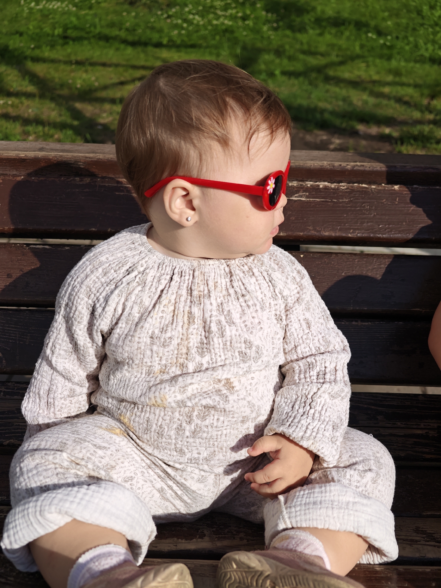 An infant wearing a patterned romper and red flower-shaped sunglasses sits on a wooden park bench. The infant's left hand rests on their lap. The background shows green grass and dappled sunlight. The scene is set in Chaikovsky, Russia.