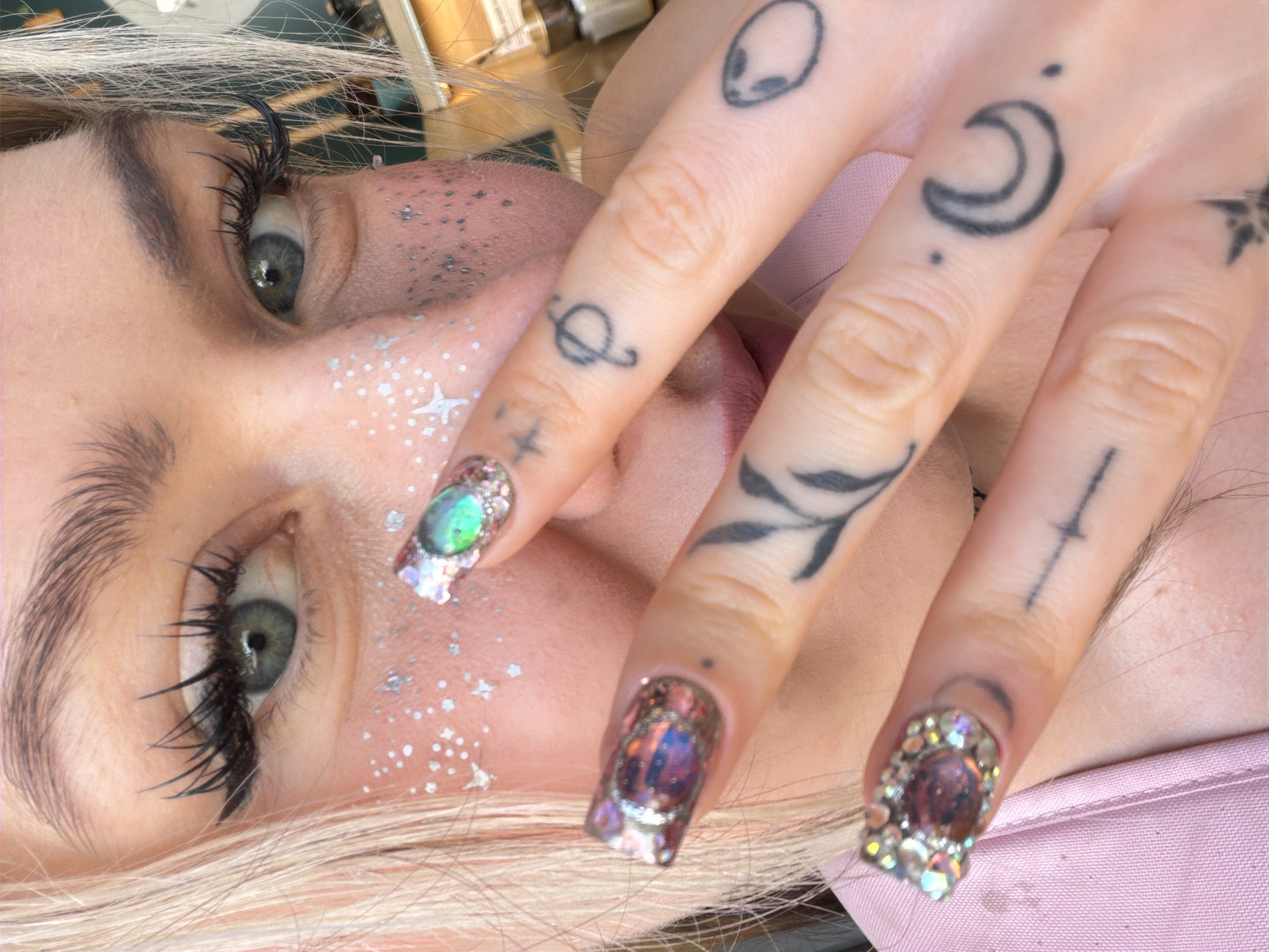 A close-up image features a woman with light blonde hair and grayish-green eyes. She is wearing heavy black false eyelashes and her face is decorated with glitter and star-shaped embellishments. Her hand, displaying several finger tattoos including an alien head, crescent moon, planet, leaves, and a sword, is positioned prominently in the foreground. Her fingernails are heavily adorned with iridescent stones. She is wearing a light pink garment. Location is not specified in image.
