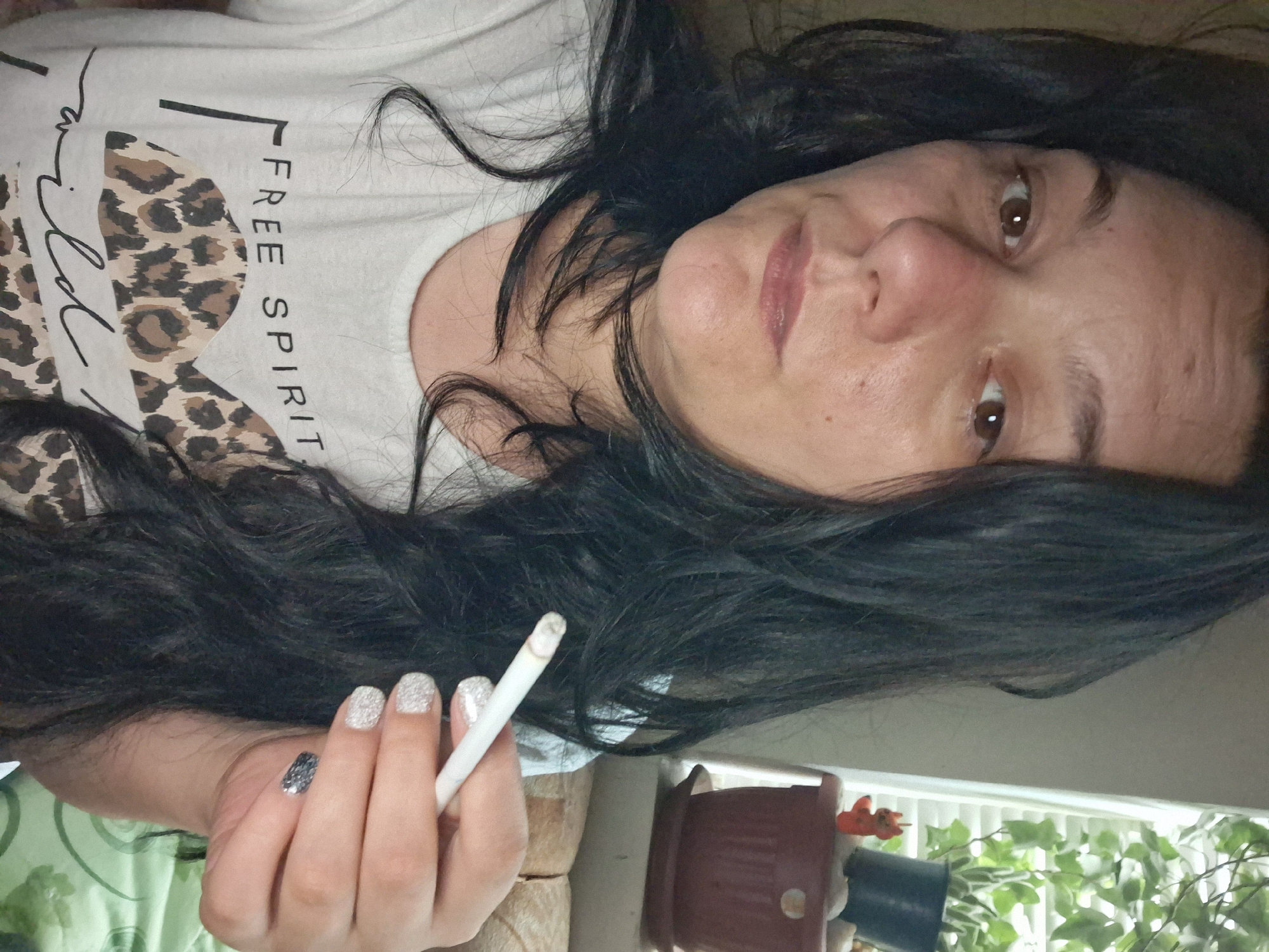 The image shows a close-up of a woman with long, dark hair, holding a cigarette in her right hand. She is wearing a white t-shirt with a graphic design that includes the words "FREE SPIRIT" and a leopard print pattern. Her fingernails are painted with a glittery polish. In the background, out of focus, is a windowsill with a potted plant and a small orange figurine. The environment appears to be indoors, possibly a residential setting. There is no discernible information in the image that allows for the specific location to be identified as Gotse Delchev, Bulgaria.
