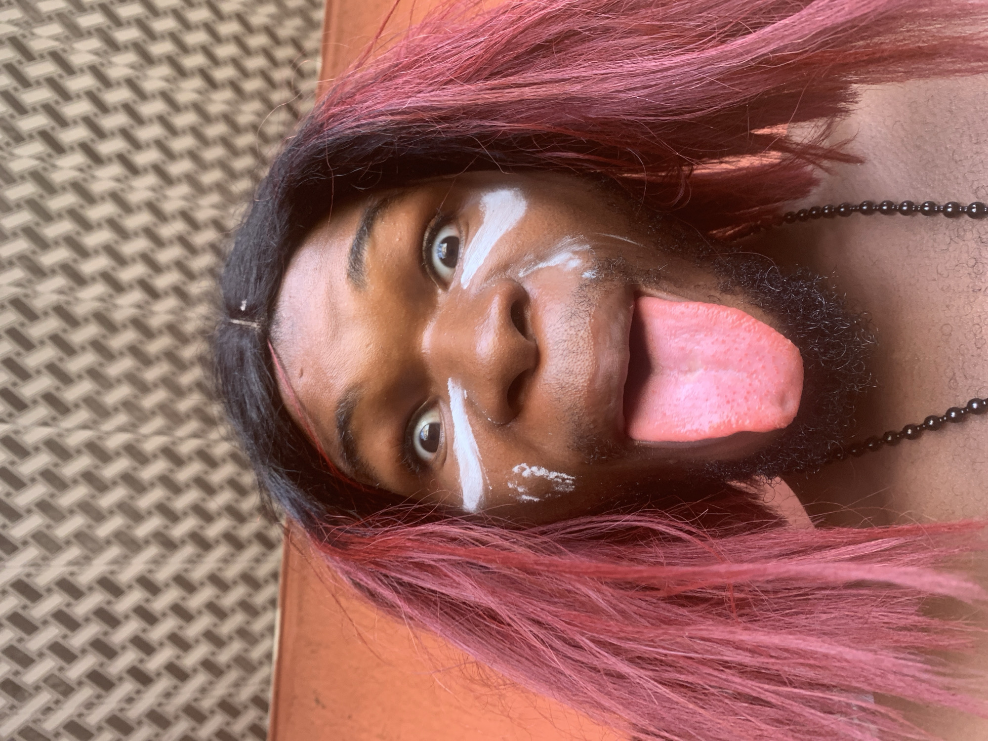 A close-up portrait features a person with dark skin and long, vibrant pinkish-red hair, oriented horizontally. The individual looks directly at the camera with wide, expressive eyes, and their tongue is playfully sticking out. White paint or makeup is applied to their face, forming a vertical stripe above one eye, a small dot on the forehead, and a smudge on the cheekbone below the other eye. A short, dark beard or stubble is visible on their chin and jawline. A dark, beaded necklace is worn around their neck. The background is composed of two main areas: a textured, brown and beige patterned surface, possibly a woven mat or floor, and a plain, solid brownish-orange surface. The lighting appears bright and even, indicating daytime. The expression conveyed is playful and direct. No text is visible in the image.