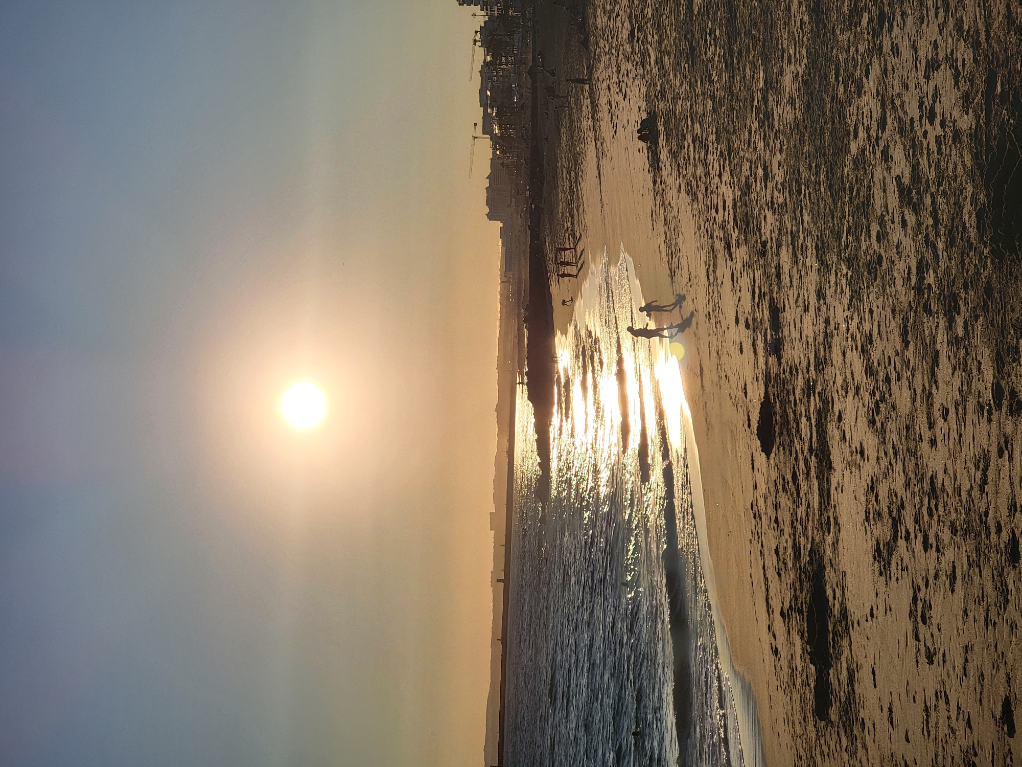 The image depicts a wide shot of a beach in Quarteira, Portugal, during sunset. The sun, a bright orb in the sky, casts a warm glow over the scene, reflecting off the gentle waves that lap the sandy shore. The ocean is a deep blue, contrasting with the golden hues of the sky and the wet sand.
Several people are visible on the beach. In the foreground, two figures, one adult and one child, walk along the water's edge, their silhouettes sharp against the bright reflection. Further down the beach, a group of individuals are gathered, some appearing to play. In the distance, the coastline is lined with buildings, indicating a coastal town. Construction cranes are visible above some of the buildings, suggesting ongoing development. The overall impression is one of a tranquil evening by the sea.