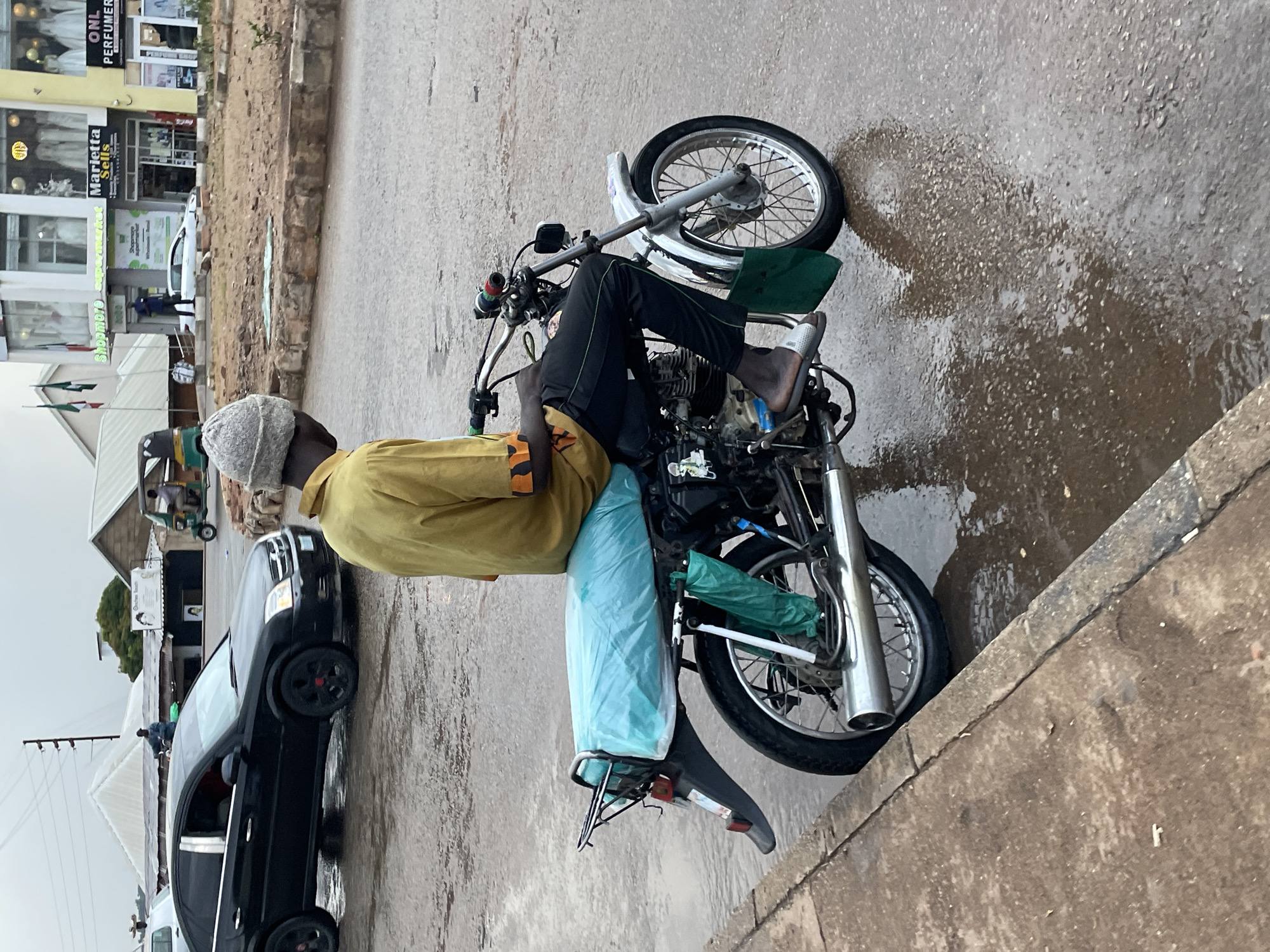 A person wearing a yellow shirt, black pants, and a grey beanie rides a motorcycle on a wet asphalt road in Jos, Nigeria. The motorcycle is carrying a large, light blue tarped object on its rear rack. The person's right foot is visible, wearing a silver slide sandal. The road is covered in shallow puddles, reflecting the overcast sky and the surrounding environment. In the background, a black sedan is parked, and several buildings with shops, including "Shopmore Supermarket," "Marietta Sells," and "ONL Perfumer," are visible. A three-wheeled auto-rickshaw is also present. The overall scene suggests a somewhat humid or rainy day.