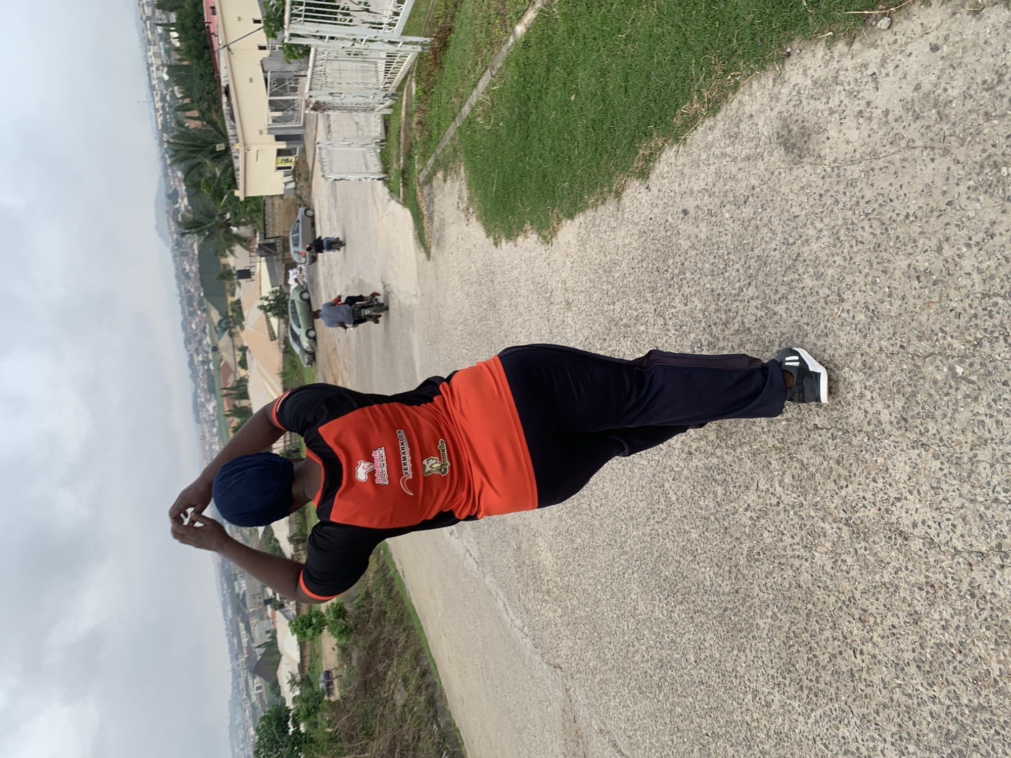 The image shows a person wearing a blue headscarf and an orange and black t-shirt, with their back to the camera, standing on a paved road. The person is raising their hands above their head. In the background, there is a cityscape with buildings and trees, indicative of a residential area. Further in the distance, a hazy skyline suggests the presence of hills or mountains. Several vehicles, including motorcycles and cars, are visible on the road and in the parking areas. The overall environment appears to be an outdoor setting within a city, possibly Abuja, Nigeria, given the architectural style of the buildings and the vegetation. The sky is overcast, suggesting a cloudy day.