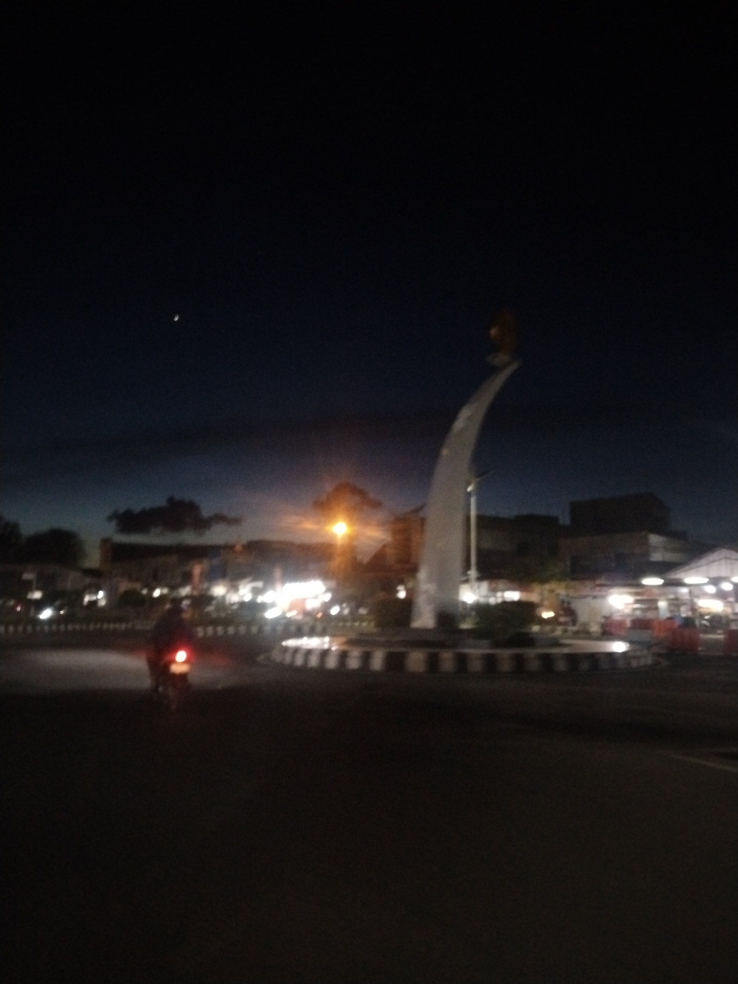 The image shows a roundabout in Lhokseumawe, Indonesia, at night. A lit monument stands in the center of the roundabout. The monument is white, curved, and topped with a gold-colored statue. A motorcycle with a red taillight is on the road approaching the roundabout. The environment includes street lights and surrounding buildings. A crescent moon is visible in the night sky.