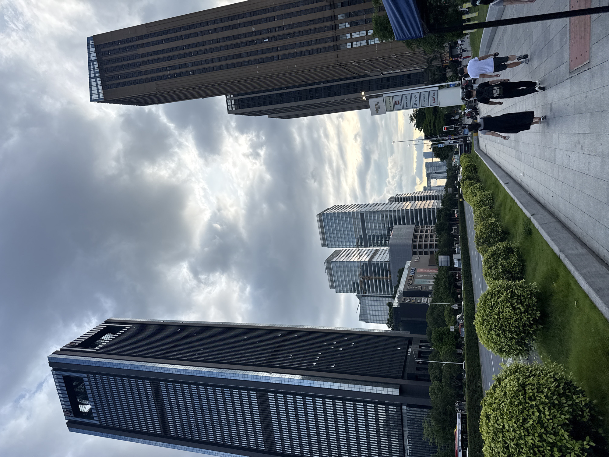 A street-level, wide-angle, low-angle shot captures a city scene in Haizhu District, China, under a cloudy sky. The perspective looks towards several tall, modern skyscrapers. The closest skyscraper on the left is a dark, rectangular building with many glass windows arranged in a grid pattern, featuring a distinctive open-top structure. To the right, a taller, brown skyscraper dominates the background, also with numerous windows. In the middle ground, other multi-story buildings, some appearing lighter and glass-fronted, are visible, with one undergoing construction.
In the foreground, a paved walkway runs parallel to a street. Several people are walking on the walkway. Three individuals are prominently featured: a woman in a black dress walking with a white bag, followed by a man in black with a backpack, and then a man in a white t-shirt and shorts. The walkway is lined with neatly trimmed, rounded green bushes and grass. The street, visible beyond the bushes, has a bus and other vehicles. A tall signpost with multiple logos is situated on the right side of the walkway. A blue awning of a structure extends into the right side of the frame. The overall lighting suggests it is late afternoon or early evening.