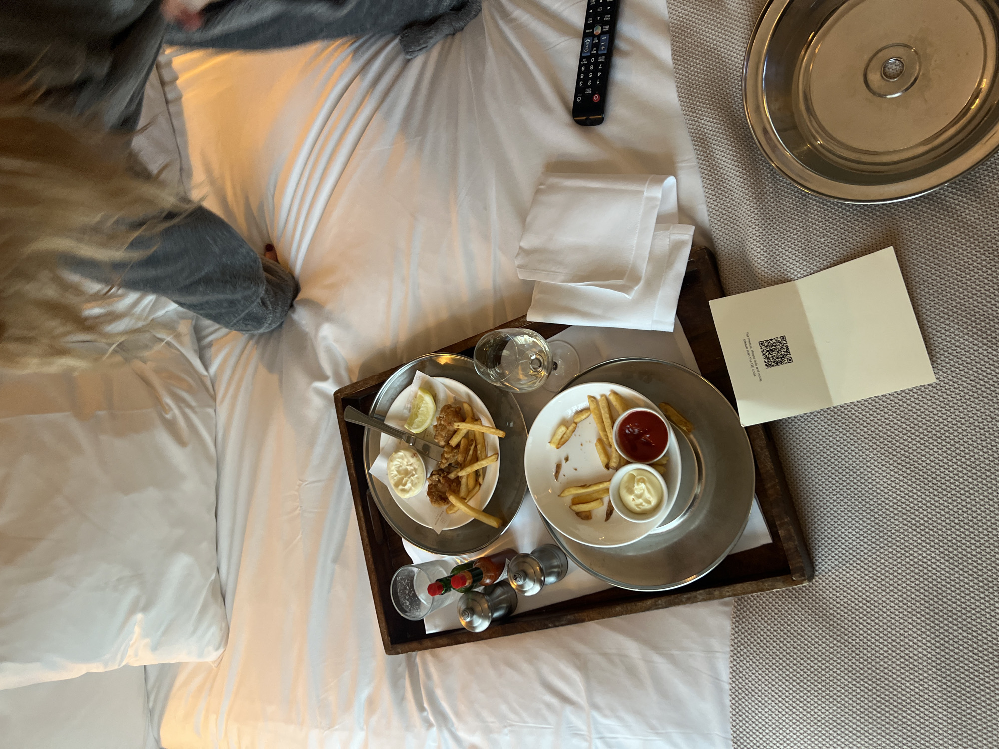 The image shows a bed in a room, likely in a hotel, with a person partially visible in the upper left. A wooden tray containing two metal plates of food, likely fish and chips, rests on the bed. A water glass, condiments, and a folded white napkin are on the tray. A hotel service menu with a QR code and a metal serving tray are also present. A remote control is visible near the head of the bed. The location is likely a hotel room, but the city of Berlin, Germany cannot be confirmed from the information in this image.