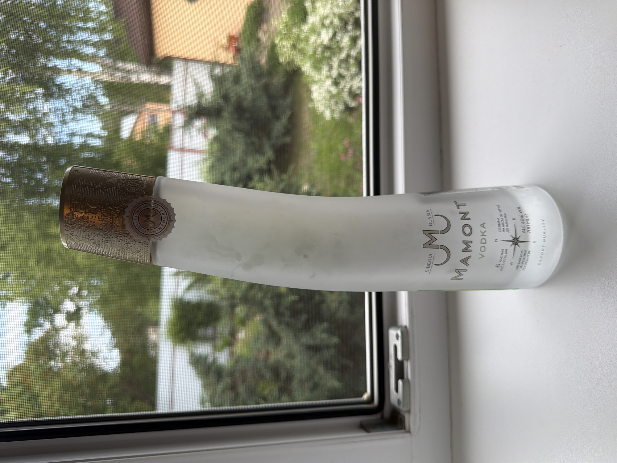 A slightly frosted, curved glass bottle of Mamont Vodka stands upright on a white windowsill. The bottle has a textured gold cap and a frosted glass body with text and graphics printed in dark grey. The background visible through the window shows a green, leafy outdoor scene with trees, bushes, and a house with an orange roof. The window is covered with a mesh screen, which distorts the background slightly. The scene appears to be an outdoor setting, possibly a backyard, seen from inside a building.