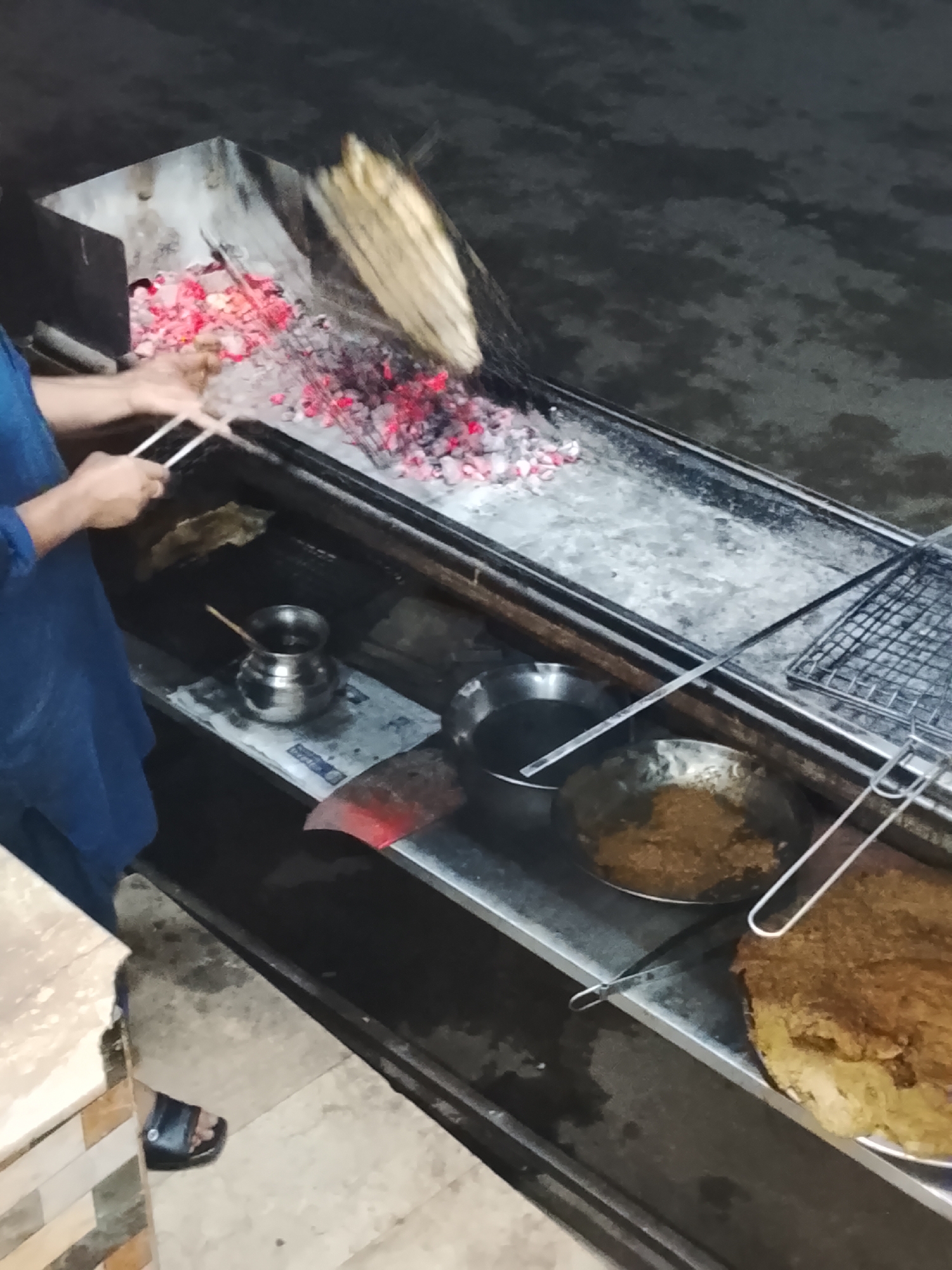 The image captures a person grilling food over hot charcoal, likely at a street food stall or an outdoor cooking establishment during nighttime. The scene is illuminated primarily by the glowing red embers of the charcoal.
A person, wearing a blue shirt or tunic and black sandals, is visible on the left side of the frame. Their hands are holding metal tongs or skewers, manipulating food items (possibly meat or vegetables) on skewers directly over the burning coals. Above the grilling area, some light-colored planks or wood pieces are in motion, possibly being used as fuel or for fanning the flames.
The grilling setup consists of a long, rectangular metal structure. On a lower shelf beneath the main grilling surface, several metal vessels are arranged. These include a small, lidded pot, a larger bowl containing a dark substance, and a flat, round dish holding a substantial amount of brown, textured food, which might be a type of bread or cooked delicacy. A piece of newspaper is placed under the small pot. To the far right, a clean metal grill rack is positioned on the counter.
The overall environment appears rustic and functional, typical of a casual eatery or a street food vendor. The dark background and the bright red glow of the charcoal confirm that it is dark outside. There are no discernible facial expressions, and any text on the newspaper is illegible.