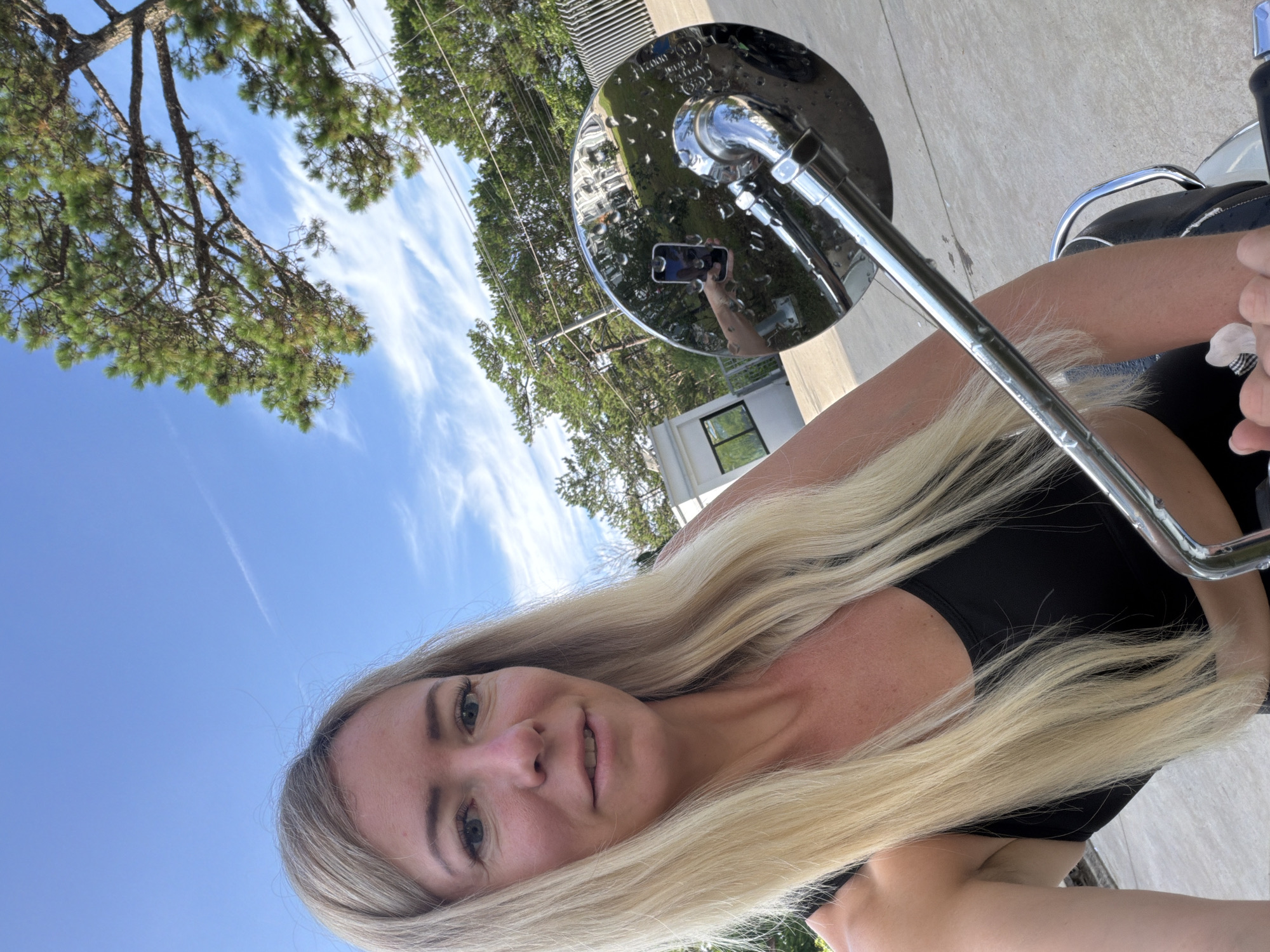In the city of Xuân Trường - Đà Lạt, Vietnam, a woman with blonde hair is taking a selfie while sitting on a motorcycle. The motorcycle's chrome rearview mirror is visible in the foreground, reflecting a portion of the scene, including a hand holding a smartphone. The woman is wearing a black tank top. Behind her, there is a bright blue sky with wispy clouds and green trees, including a large pine tree. In the background, a modern white building with windows and a fence can be seen. The ground is paved with light-colored concrete.