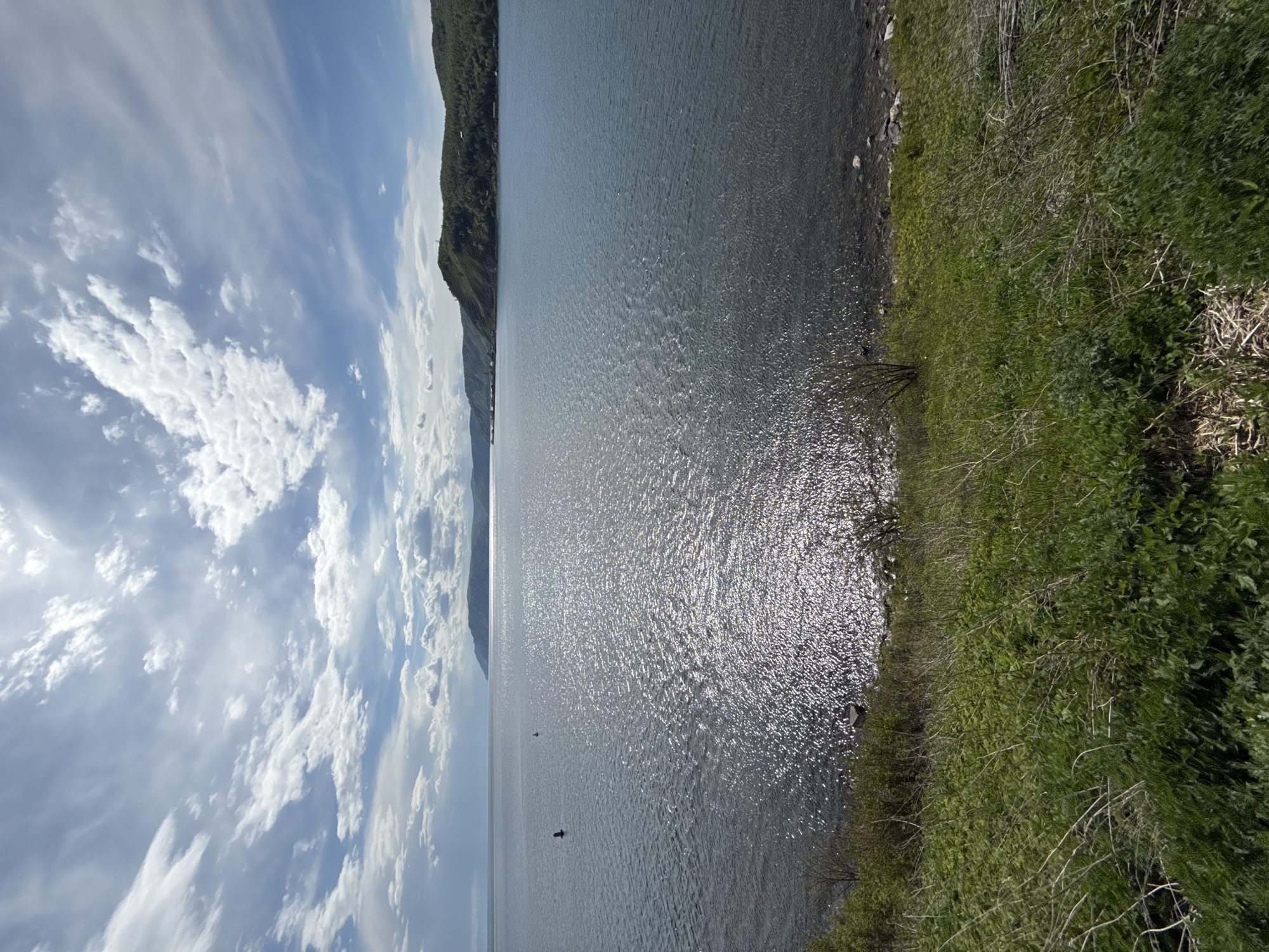 The image shows a body of water, possibly Lake Baikal, in Listvyanka, Russia, reflecting sunlight, bordered by a shoreline with vegetation. A mountain range is visible in the background with a cloudy sky.