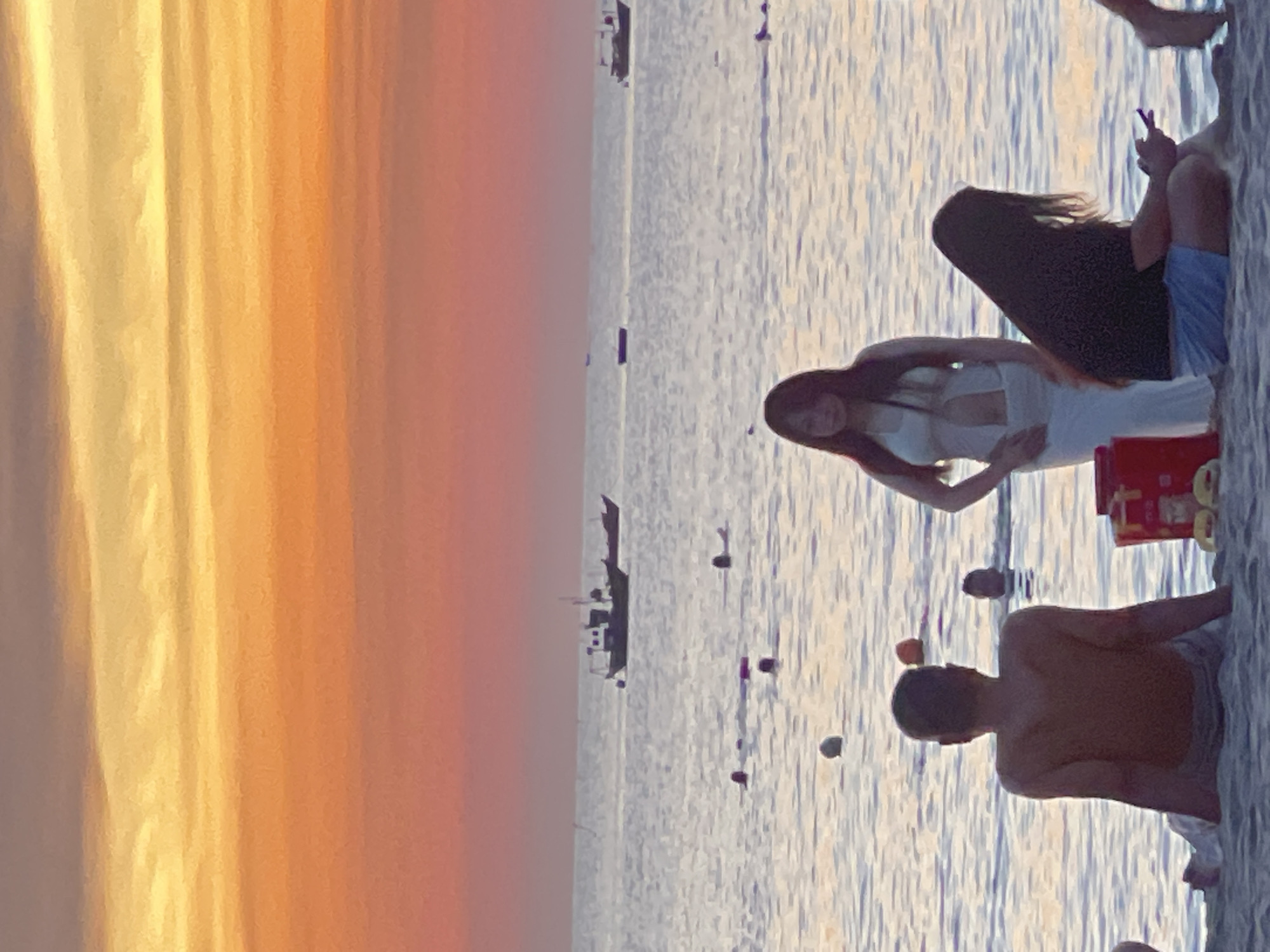 A group of people are on a beach in Da Nang, Vietnam, at sunset. The sky is a vibrant mix of orange and yellow. In the foreground, a man sits on the sand facing away from the camera, with his back to the ocean. To his right, a woman in a white dress stands facing the camera, looking slightly to her left. Next to her, another person with long dark hair sits on the sand, looking down at a phone in their hands. In the water, several people are swimming, and in the distance, two boats are visible on the horizon. The overall scene depicts a relaxed evening on the beach.