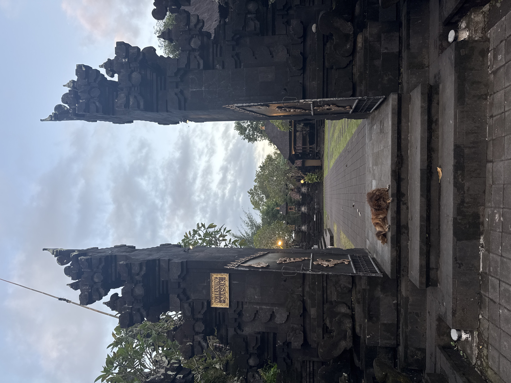 The image shows an ornate, dark stone gateway in Dawan, Indonesia. The gateway is flanked by two intricately carved pillars. A sign with text in a foreign language is mounted on the left pillar. Beyond the gateway, a paved path leads to a courtyard with traditional Indonesian architecture, including thatched-roof buildings and stone statues. A shaggy, brown dog is lying down on the stone steps directly in front of the gateway. The sky above is cloudy with hints of twilight. The overall scene evokes a sense of tranquility and ancient cultural heritage.