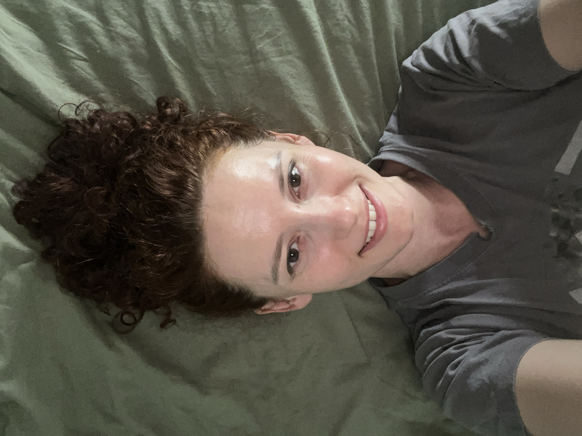 A selfie-style image shows a woman lying on a green bedsheet. She has curly, reddish-brown hair and fair skin. She is wearing a gray t-shirt and is smiling. The background consists solely of the wrinkled green bedsheet. There is no indication of the location being Saint Petersburg, Russia or any other identifiable place.