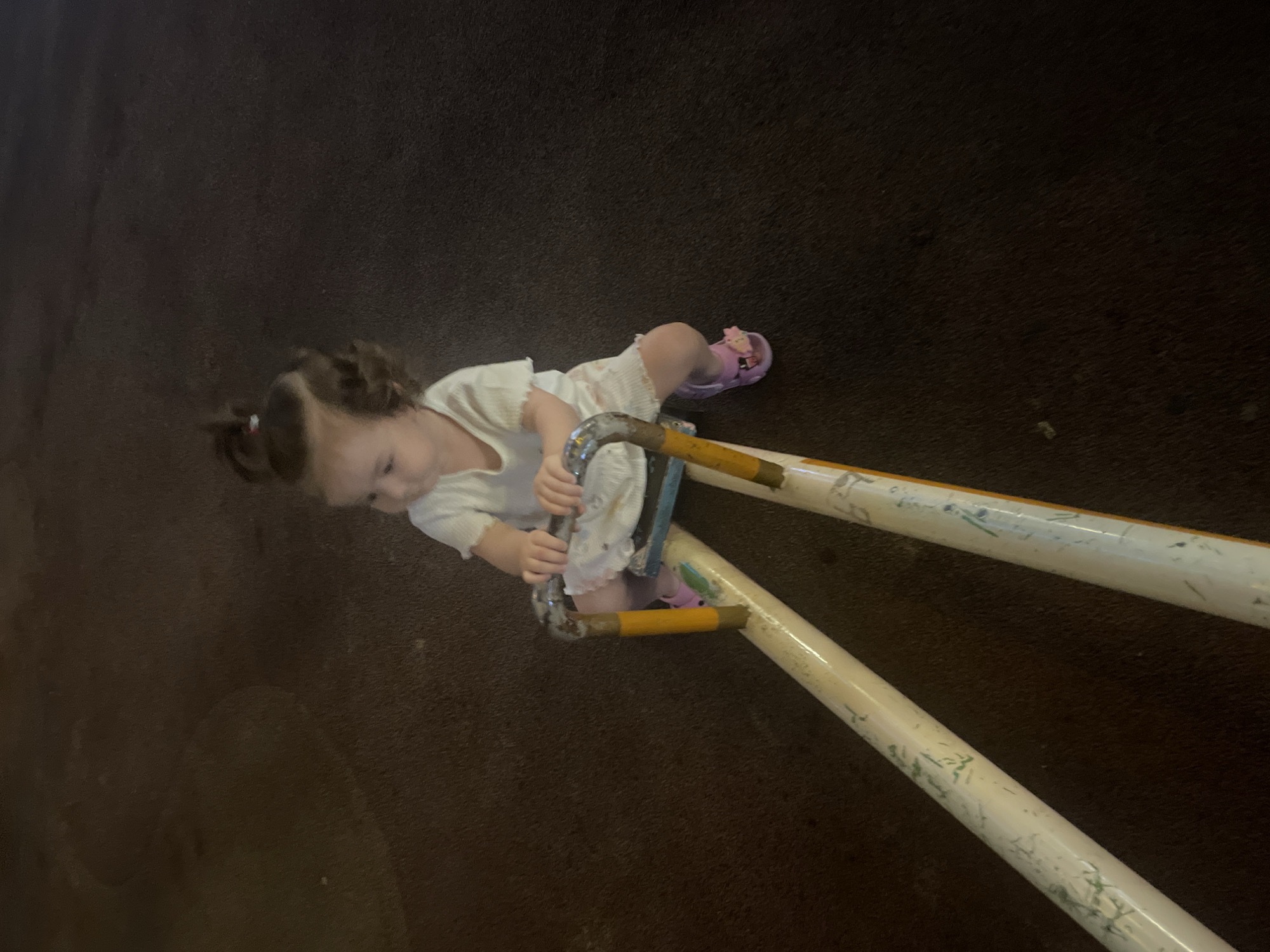 A young child with curly brown hair is sitting on a seesaw in a dimly lit outdoor area. The child is wearing a white t-shirt and white shorts, with purple sandals on their feet. The seesaw appears to be made of metal and has yellow accents. The ground is a dark, textured surface, possibly asphalt or rubberized playground material. The lighting suggests it is nighttime, possibly in a park or playground in Dushanbe, Tajikistan. The child is holding onto the handles of the seesaw and looking to the left.