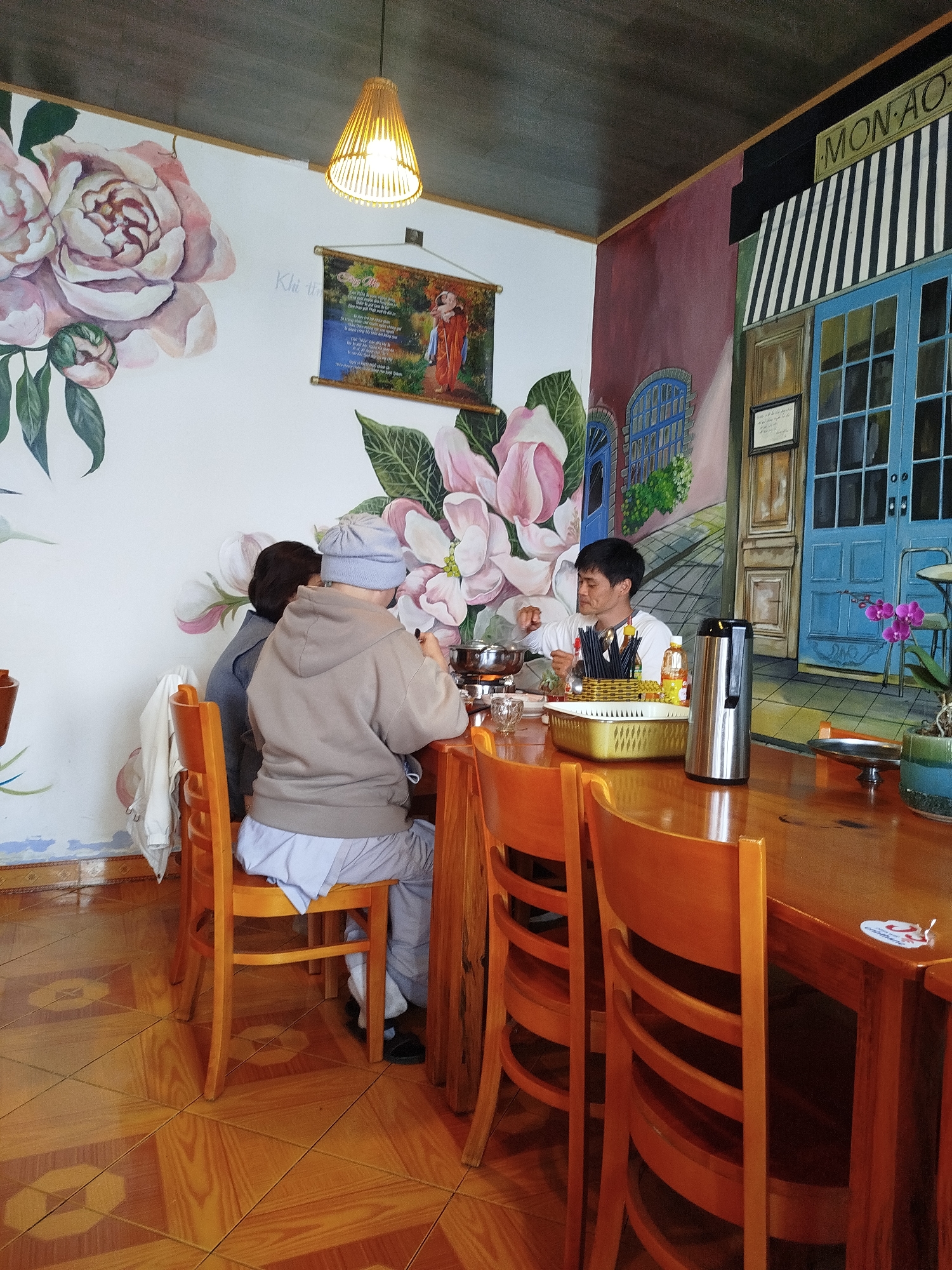 Two people are seated at a wooden table in what appears to be a restaurant in Da Lat, Vietnam. A man sits across from them at the same table. They are near a wall with a floral painting and a painting that resembles a store front. All are sitting in matching wooden chairs. The table has a pot for cooking, utensils and other items. The floor is tiled. A light hangs above the table.