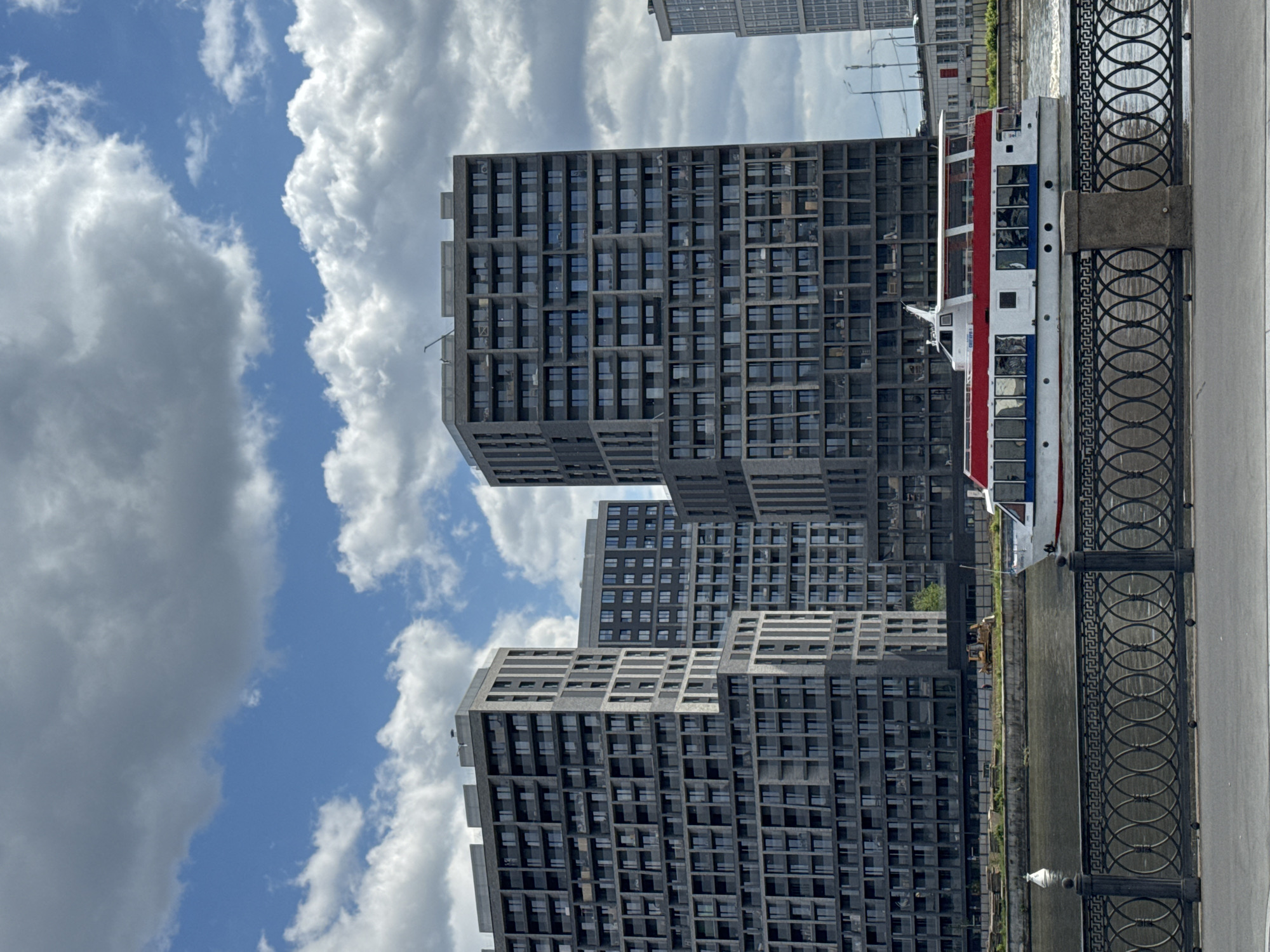 The image presents an urban waterside scene in Moscow, Russia, captured during daytime under a partly cloudy sky. The perspective is from an embankment, looking across a river or canal towards modern architecture.
In the immediate foreground, a dark, ornate metal fence with circular and geometric patterns runs along a paved area, bordering the water. The river appears calm, reflecting the ambient light.
Mid-ground features a white passenger boat with a prominent red stripe along its upper deck and a narrower blue stripe below. The boat has numerous windows along its side, and discernible text on its hull reads "ВОЛГА" (Volga), indicating its name or connection to the Volga River, possibly serving as a river cruise vessel.
The background is dominated by a cluster of tall, contemporary buildings. These structures exhibit a distinctive, grid-like facade with numerous rectangular windows set within a dark, possibly concrete or stone, framework. The buildings vary in height and design, some appearing to be terraced or stepped, contributing to a modern urban skyline.
The sky above is a blend of clear blue patches and large, billowy white and grey clouds. No specific people are clearly identifiable, and the overall scene conveys the general atmosphere of a bustling city waterfront.