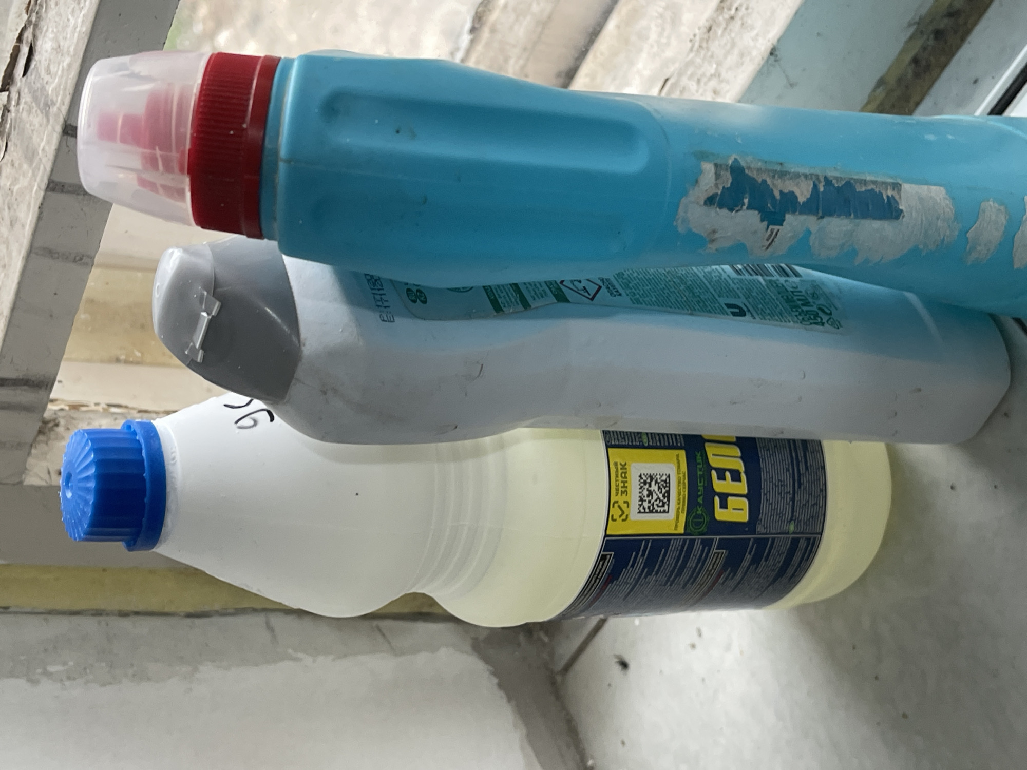 This media file displays three bottles of cleaning supplies stacked horizontally. The topmost bottle is a bright blue plastic bottle with a red screw-on cap, partially covered by a clear plastic dispenser. The middle bottle is a light gray plastic bottle with a gray cap. The bottommost bottle is a white plastic bottle with a blue screw-on cap. This bottle is filled with a pale yellow liquid. The label on the bottom bottle is visible and features a QR code, along with text in Cyrillic. Some text visible on the label includes "ЧЕСТНЫЙ ЗНАК" and "БЕЛИЗНА". The bottles appear to be resting on a ledge or surface near a window. The background suggests an indoor setting, possibly a window sill, with some outdoor elements visible through what might be a window or open space. The lighting suggests daytime. The overall scene is static, with no active event taking place.