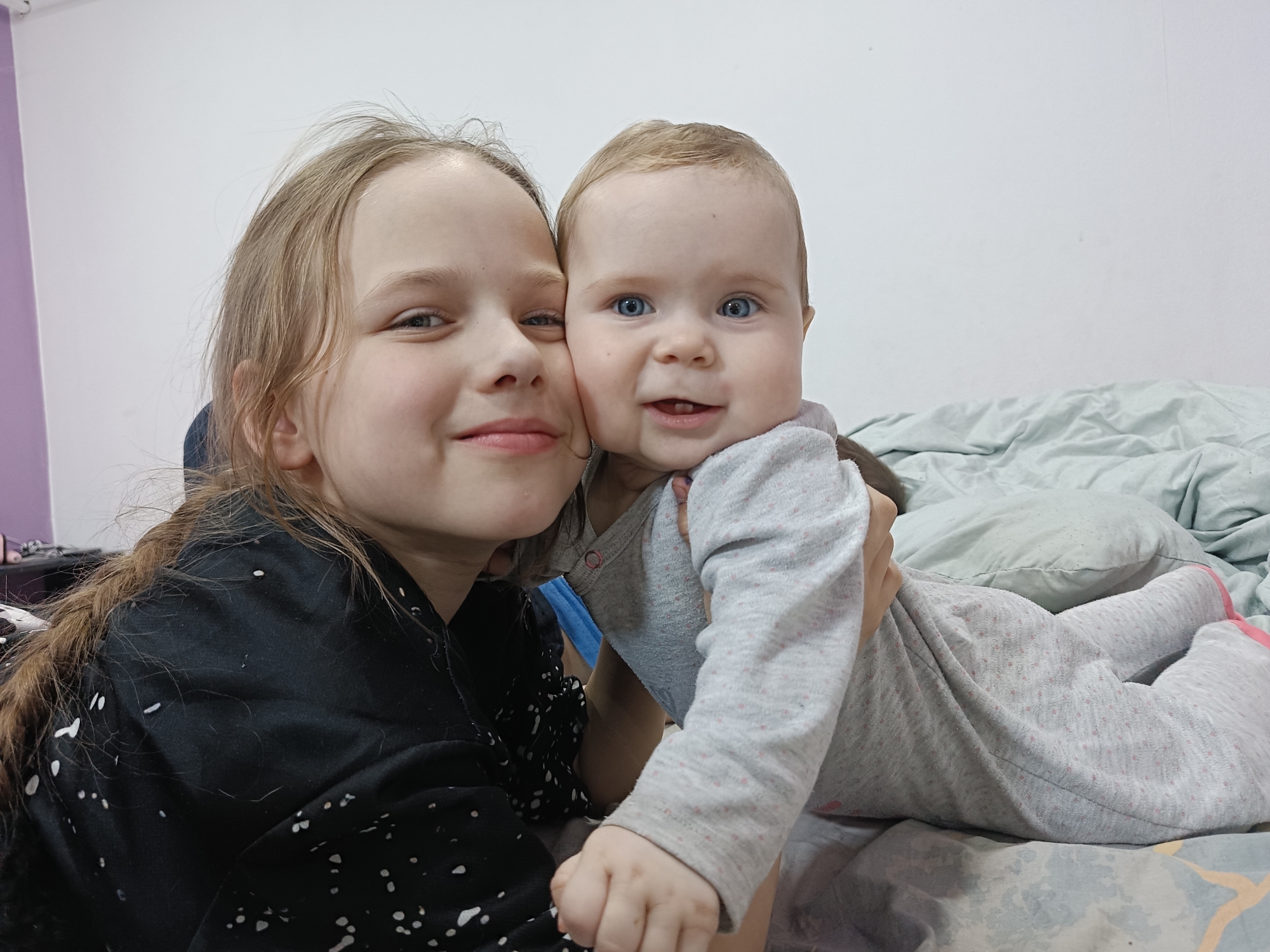 Two light-skinned individuals, one a child and one an infant, are positioned close together, likely indoors. The child, estimated to be around 8-10 years old, is on the left side of the frame. She has blonde hair styled in a braid that falls down her left shoulder, fair skin, and a light-colored complexion. She is wearing a black, long-sleeved shirt with white spots or embellishments. She is smiling, and her face is positioned near the infant's face.
The infant, on the right, is wearing a grey, long-sleeved onesie with light-pink dots and pink accents at the foot. The infant is lying on a bed covered with a blue-and-grey patterned bedspread and appears to be held by the child. The infant also has fair skin, blue eyes, and light brown hair.
The background consists of a plain white wall. The lighting is even, and the scene appears to be a casual, candid snapshot, perhaps taken with a smartphone. The specific location within the city of Rusalevka, Russia cannot be determined from the provided image.
